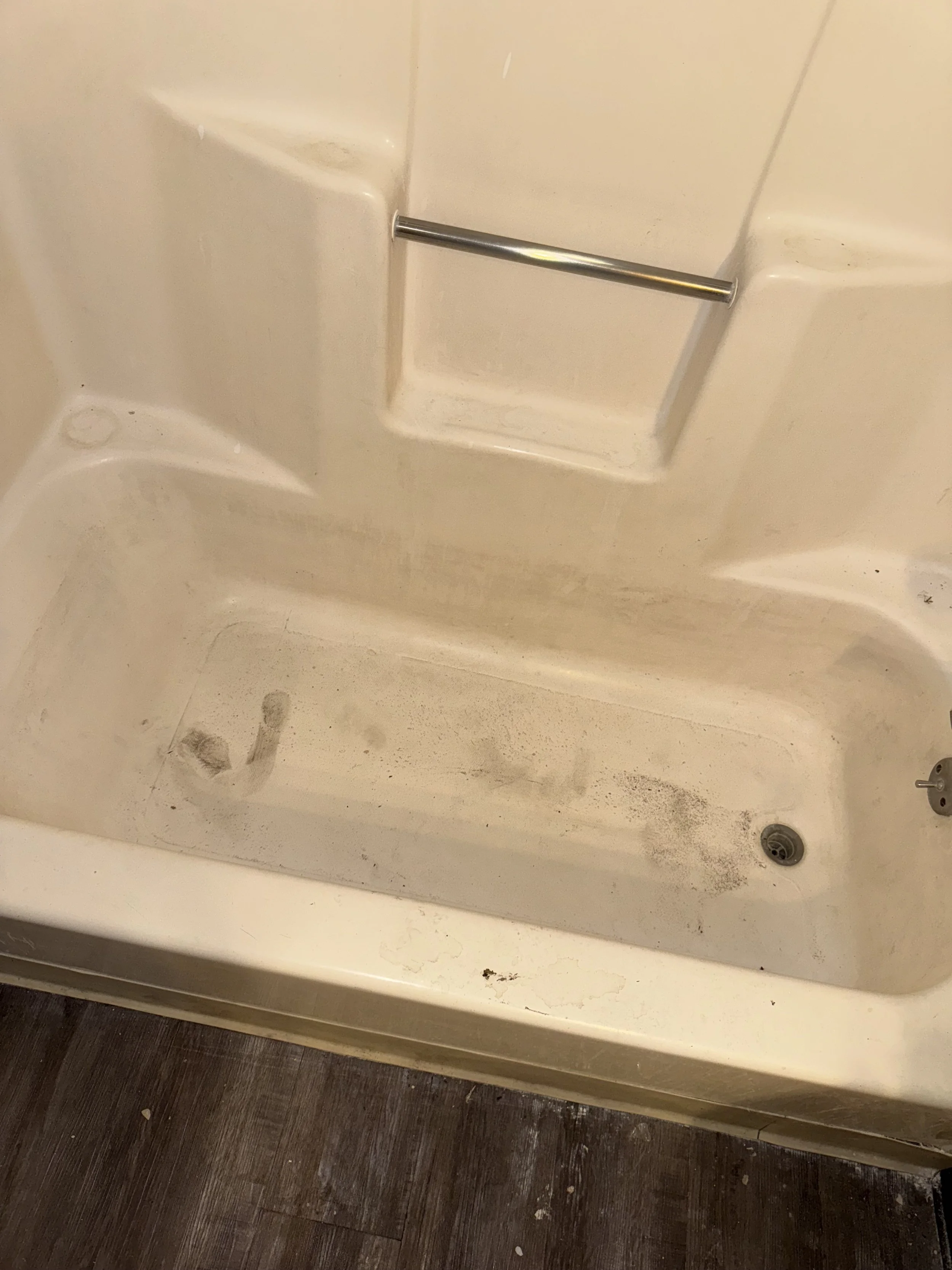 Dirty bathtub with visible dirt, scuff marks, and stains, with a metal grab bar on the wall and a drain at the bottom, situated above dark wood flooring.