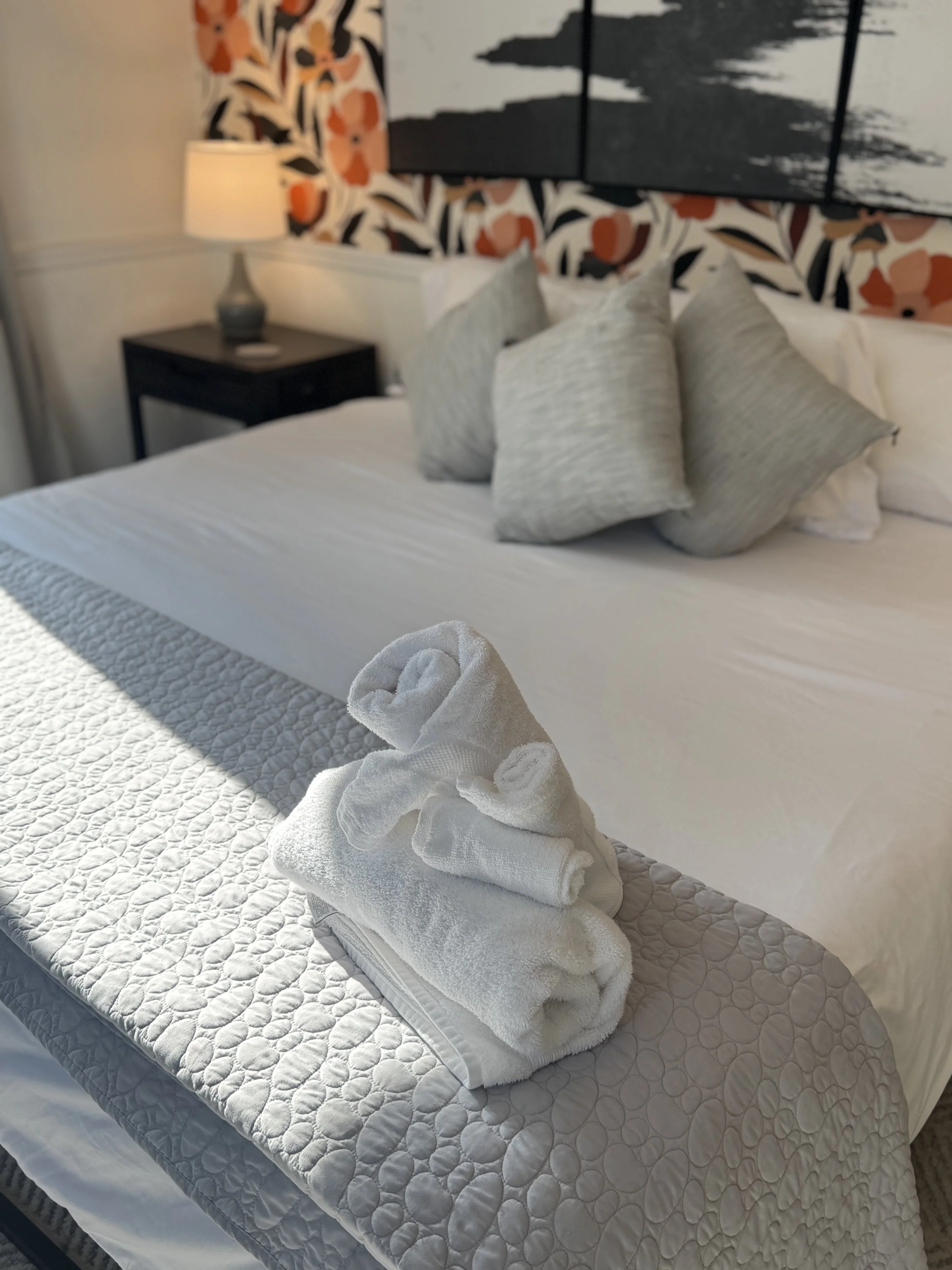 A neatly made bed with white linens, a quilted gray bed runner, and gray pillows. Towels are folded and shaped into a swan and a smaller towel on the bed. A bedside table with a lamp is visible, and there is decorative floral wallpaper and artwork behind the bed.