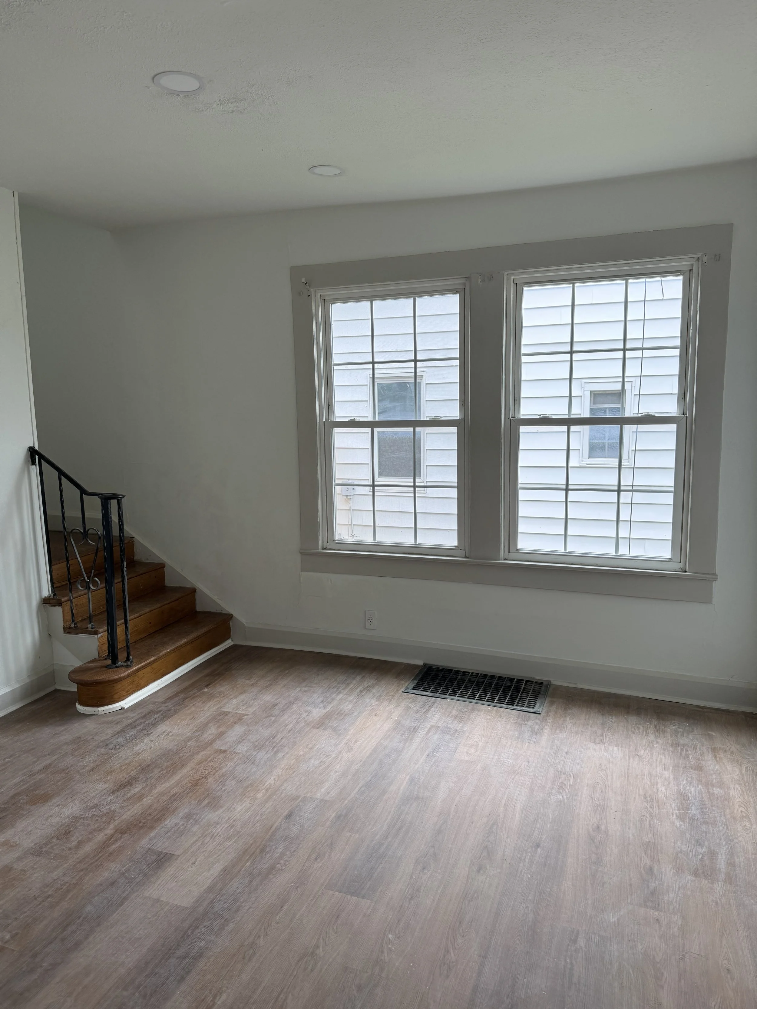 Empty living room with two large windows, wooden floor, and a small staircase with a black metal railing leading up to another area.