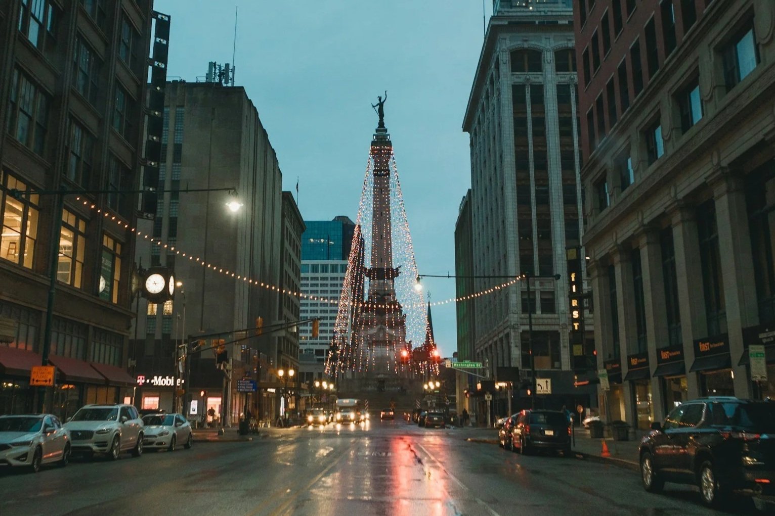 City street at dusk with a decorated monument in the center, string lights overhead, parked cars along the sides, and tall buildings surrounding the scene.