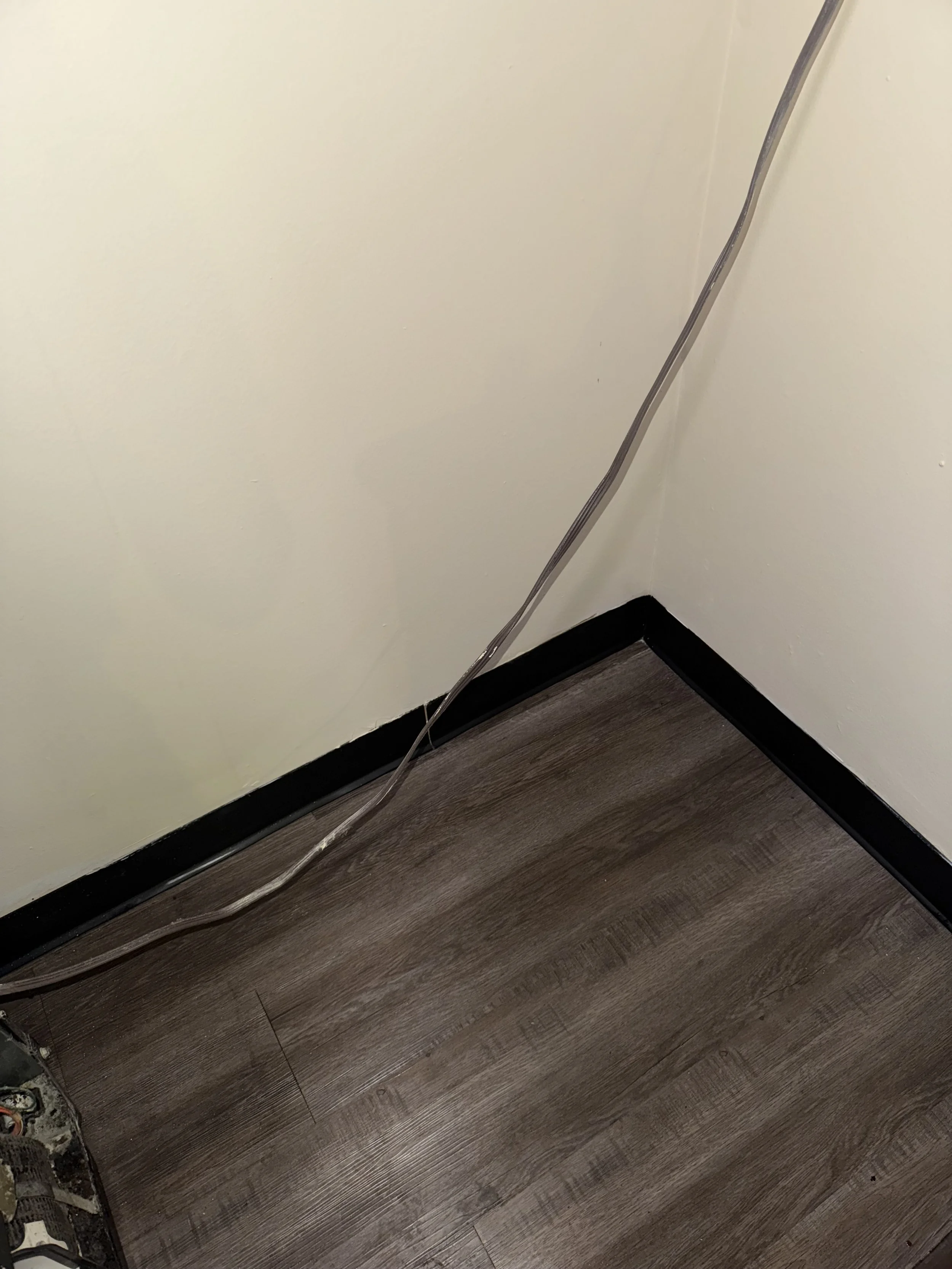 Close-up of a corner with electrical wires running along the wall and floor, and a black baseboard.
