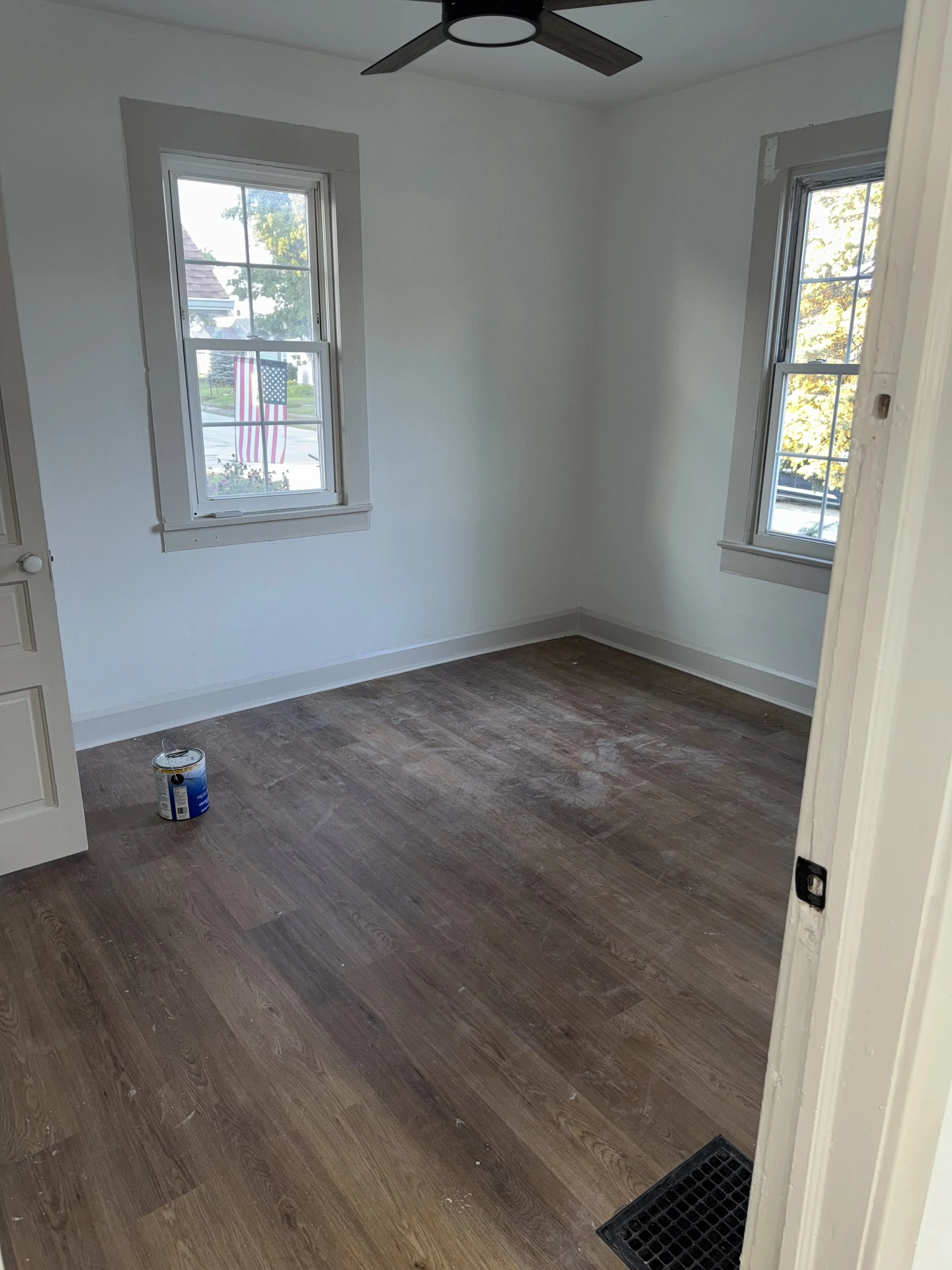 Empty room with two windows, white walls, brown hardwood floor, a ceiling fan, and a can of paint on the floor.