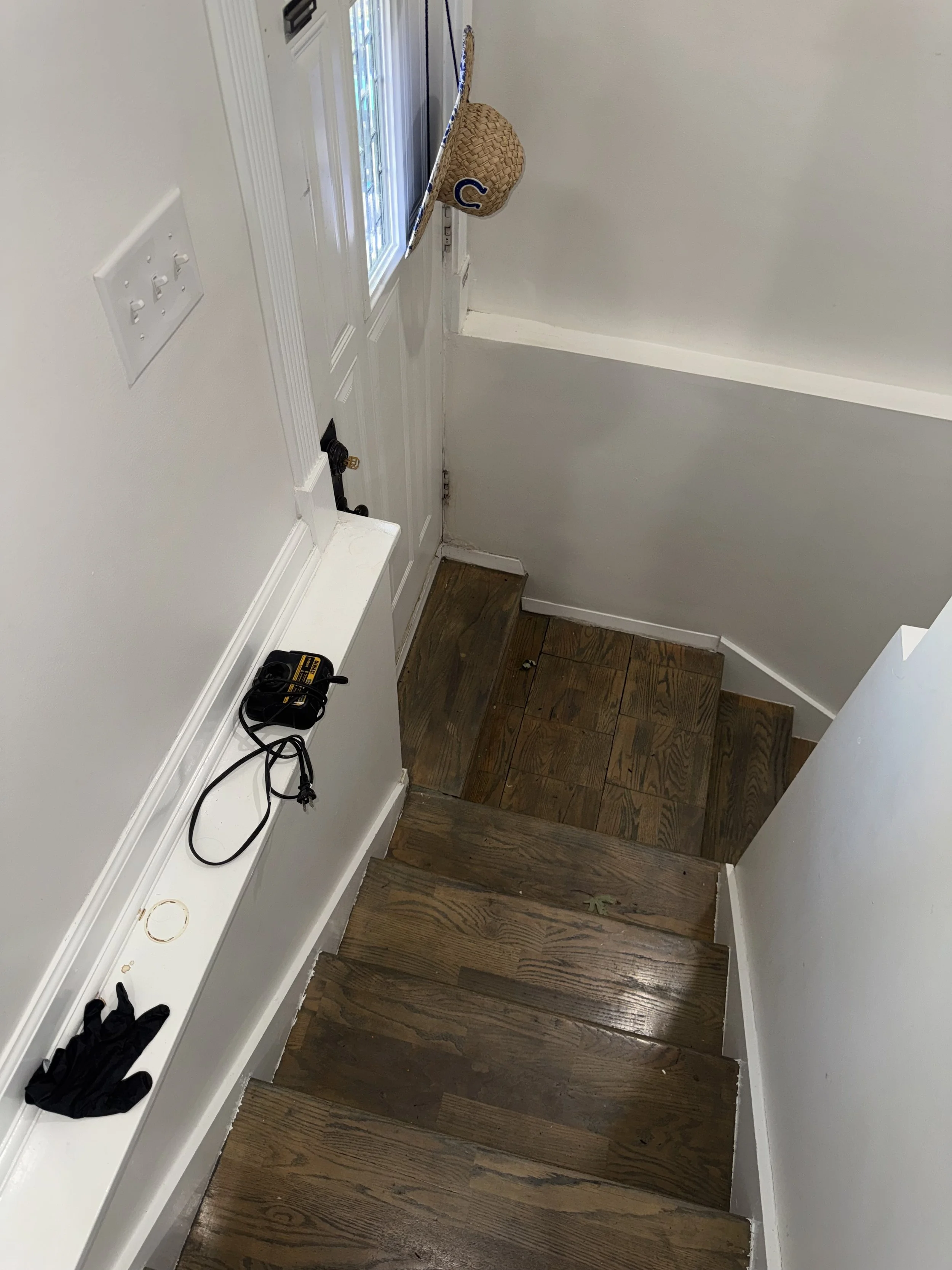 Interior of a home entrance with wooden stairs, a white wall, a door with a window, a hat hanging on the door, a small round seal on the wall, black gloves, and a black extension cord on the white shelf.