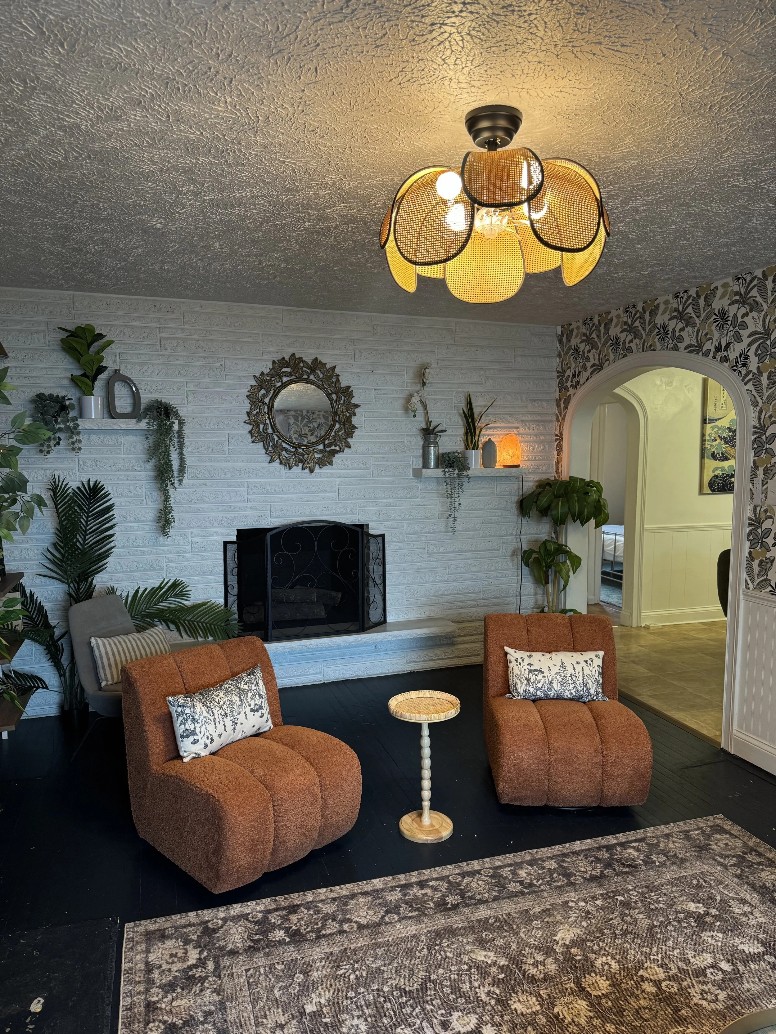 Living room with orange chairs, white brick fireplace, decorative mirror, plants, shelf with decor, and a unique yellow flower ceiling light fixture.