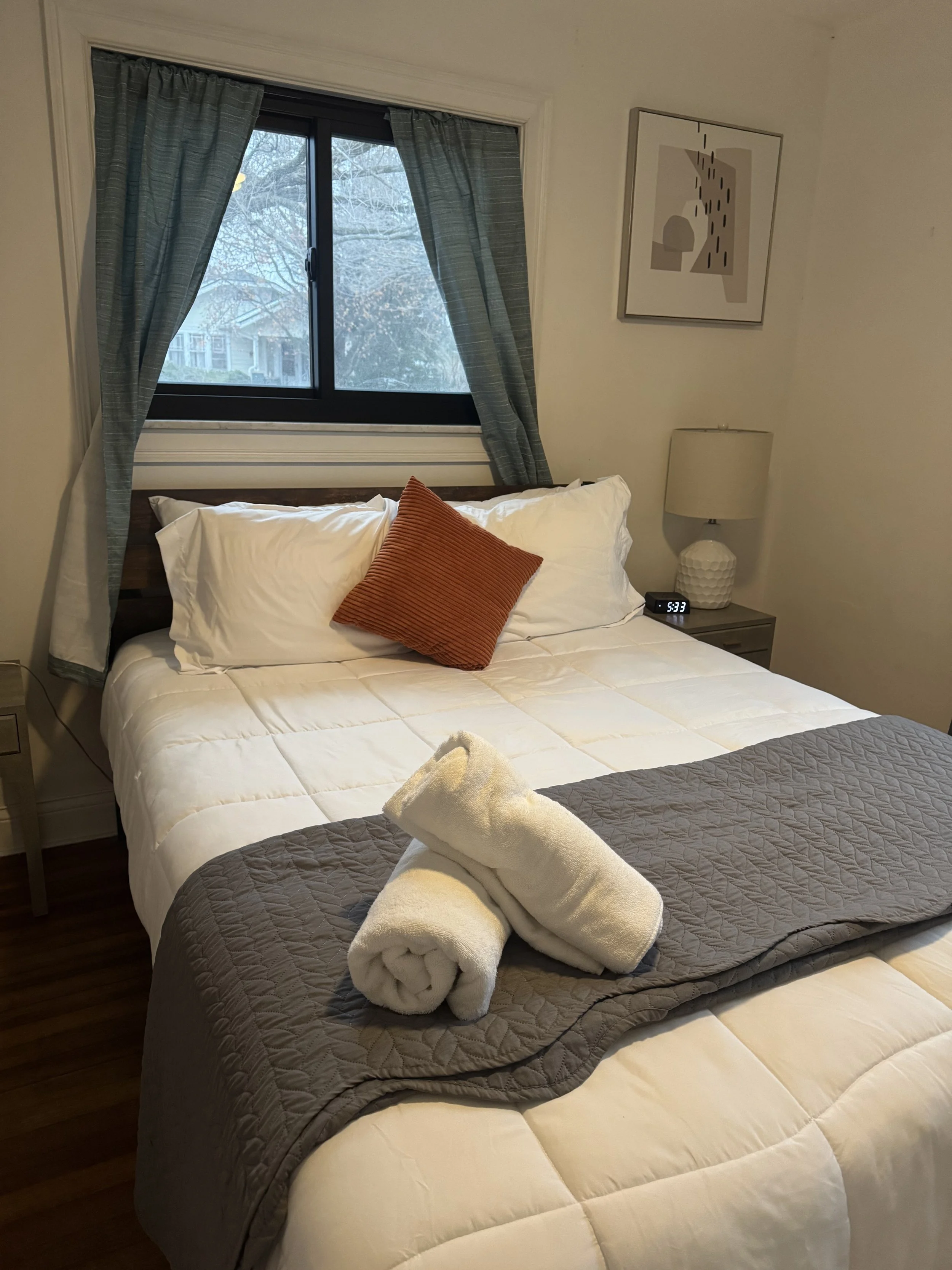 A neatly made bed with white pillows, an orange decorative pillow, and rolled white towels at the foot, in a bedroom with a window, curtain, nightstand, and lamp.