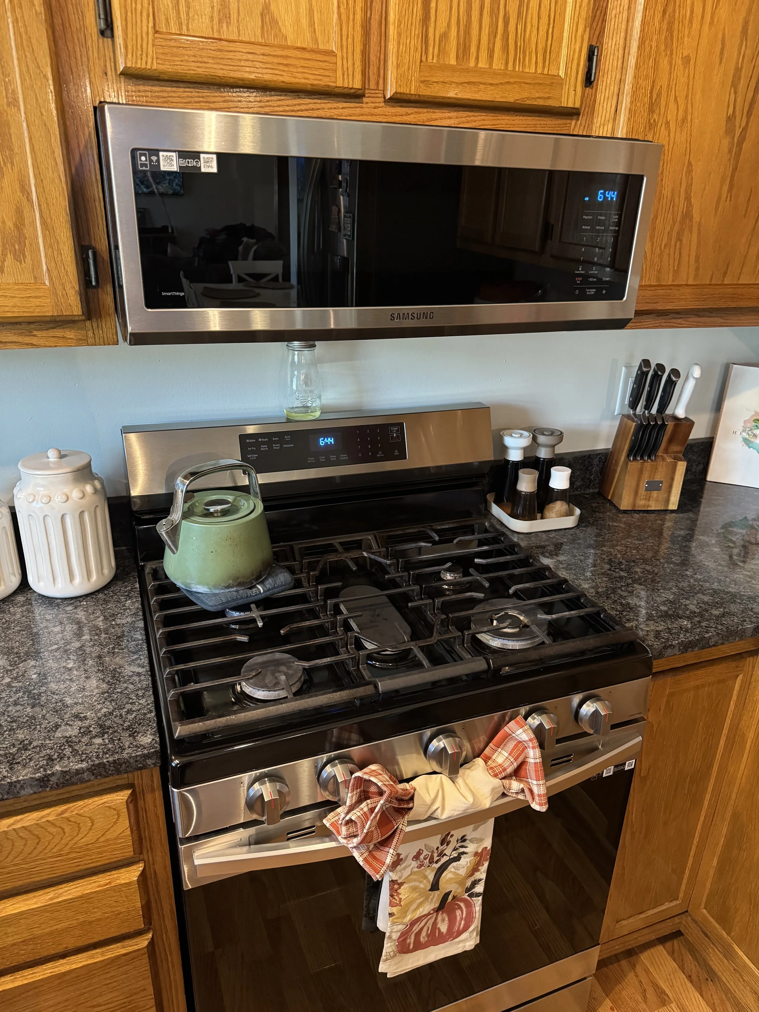 Stainless steel microwave mounted above a black gas stove in a kitchen with wooden cabinets and a black granite countertop, with a green teapot on the stove and various kitchen utensils nearby.