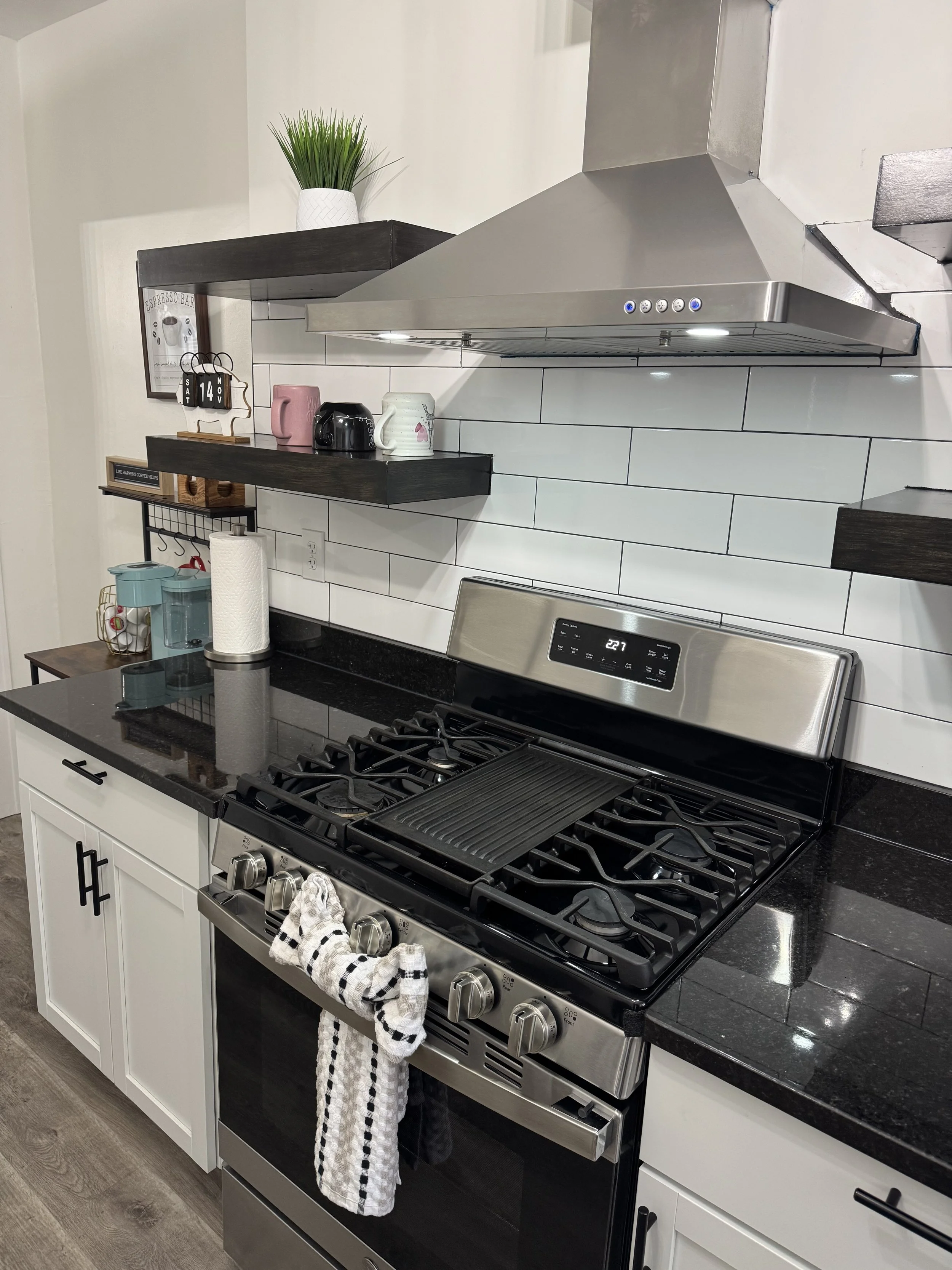 Modern kitchen with a black granite countertop, stainless steel stove with oven, white cabinets, black handles, black soapstone backsplash, stainless steel range hood, two open wooden shelves with mugs, paper towels, decorative items, and a potted plant on top.