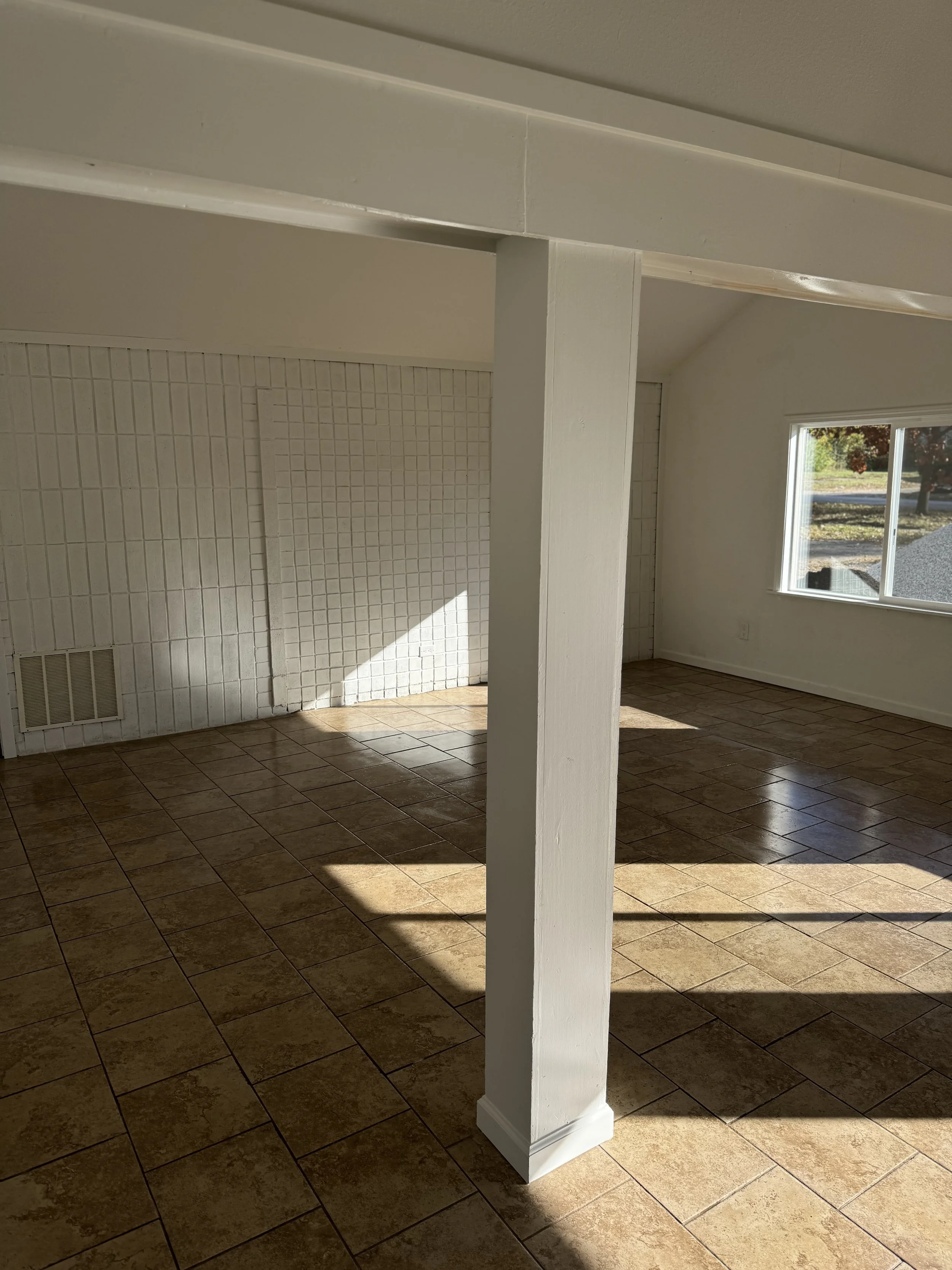 Empty room with tile flooring, white painted pole in the center, white textured wall on the left, window on the right side, sunlight casting shadows on the floor.