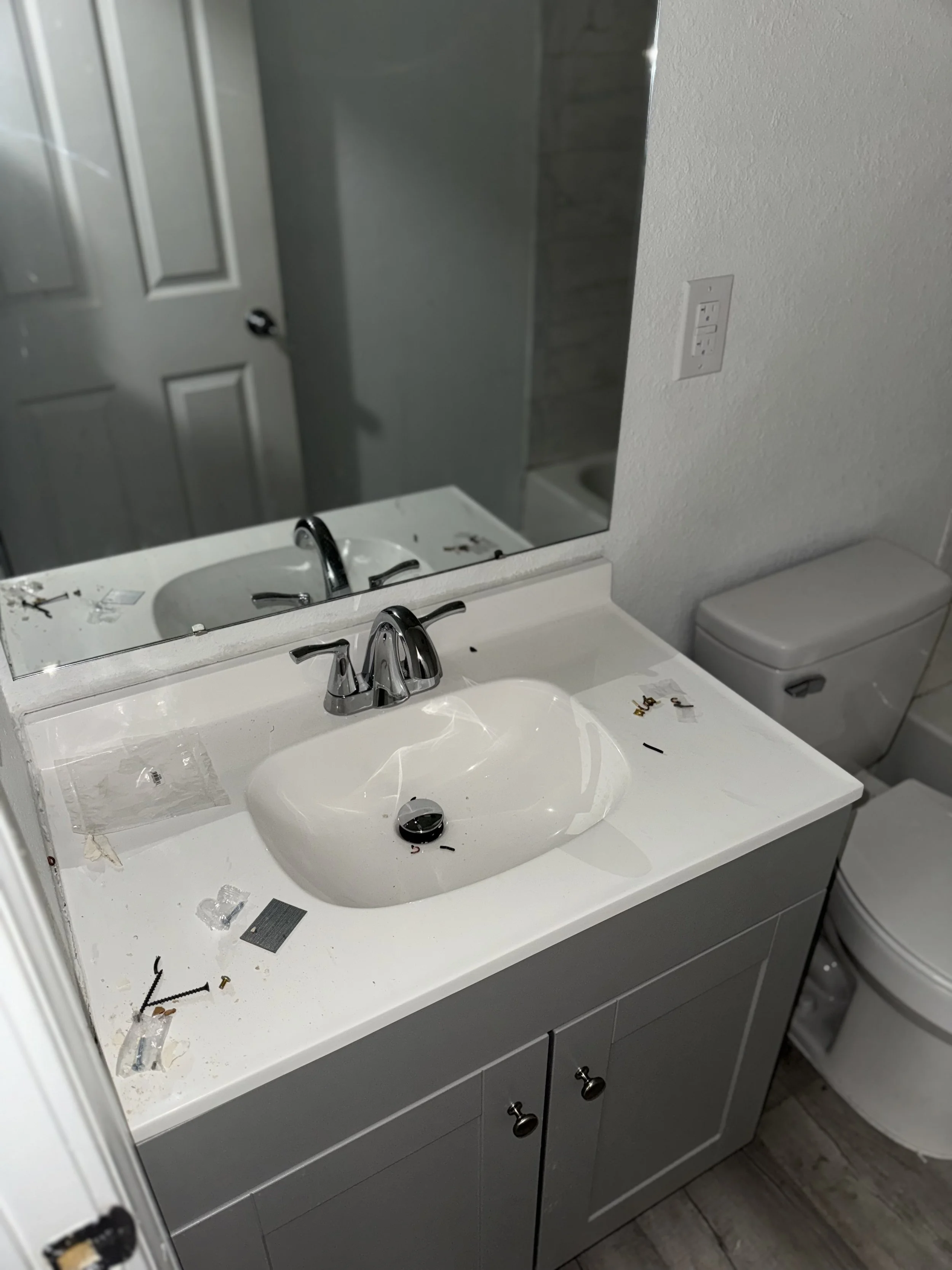 Bathroom with a white sink and gray cabinet, cluttered with debris, a mirror, a white toilet, and a textured wall.