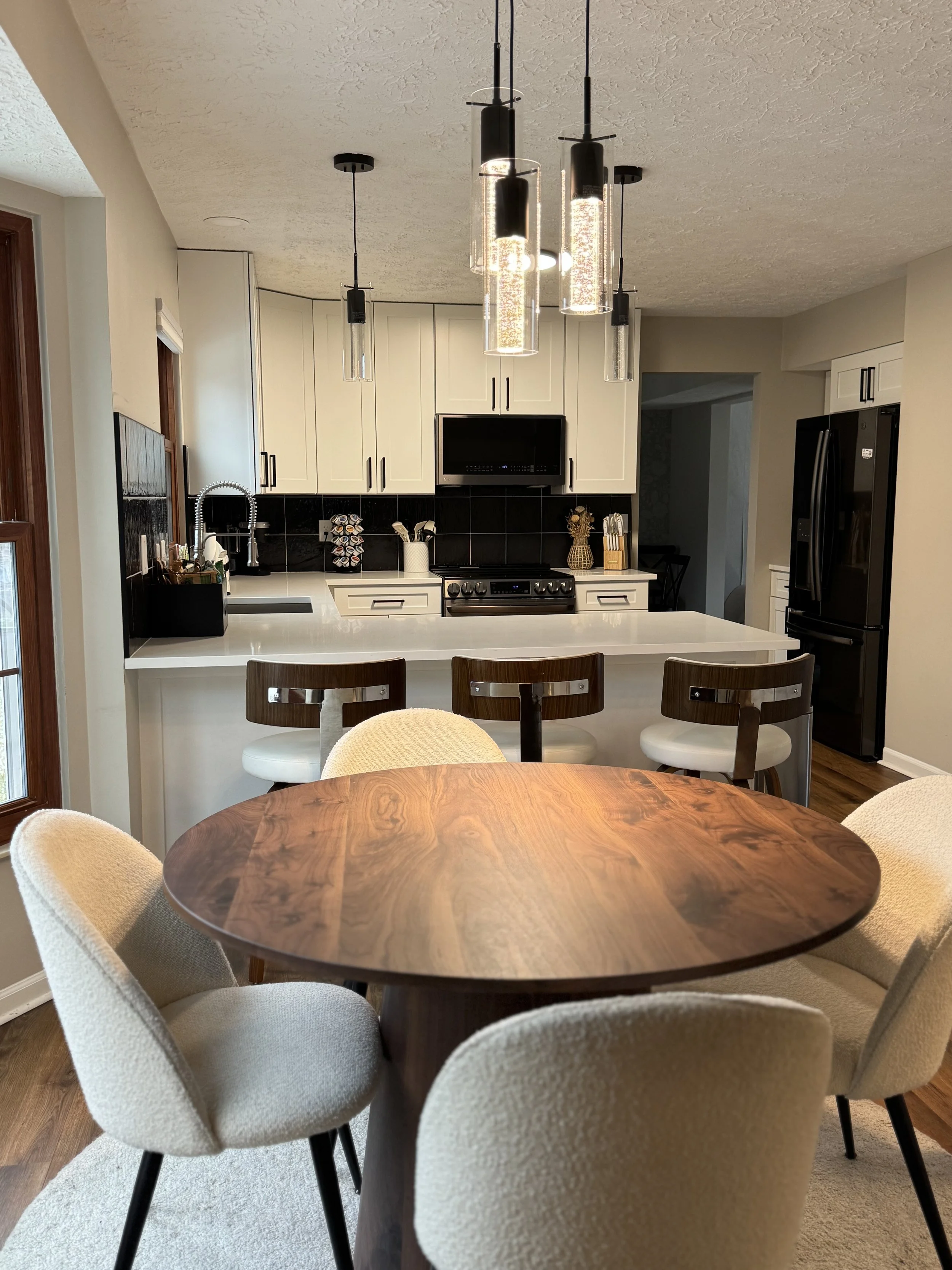 Kitchen with white cabinets, black backsplash, black appliances, and a white countertop island. A round wooden dining table with cream upholstered chairs is in the foreground, and pendant lights hang from the ceiling.