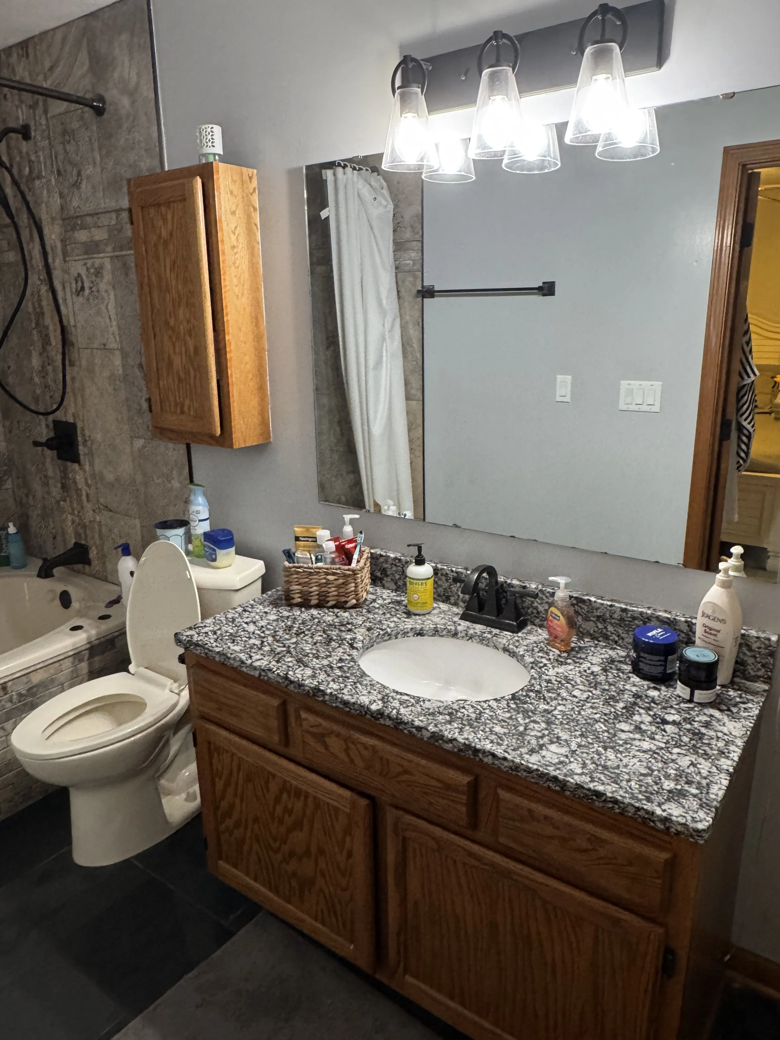Bathroom with granite countertop sink, wooden cabinet, large mirror, and light fixture with three glass shades. Toilet and bathtub are visible, along with various toiletries and a basket on the counter.