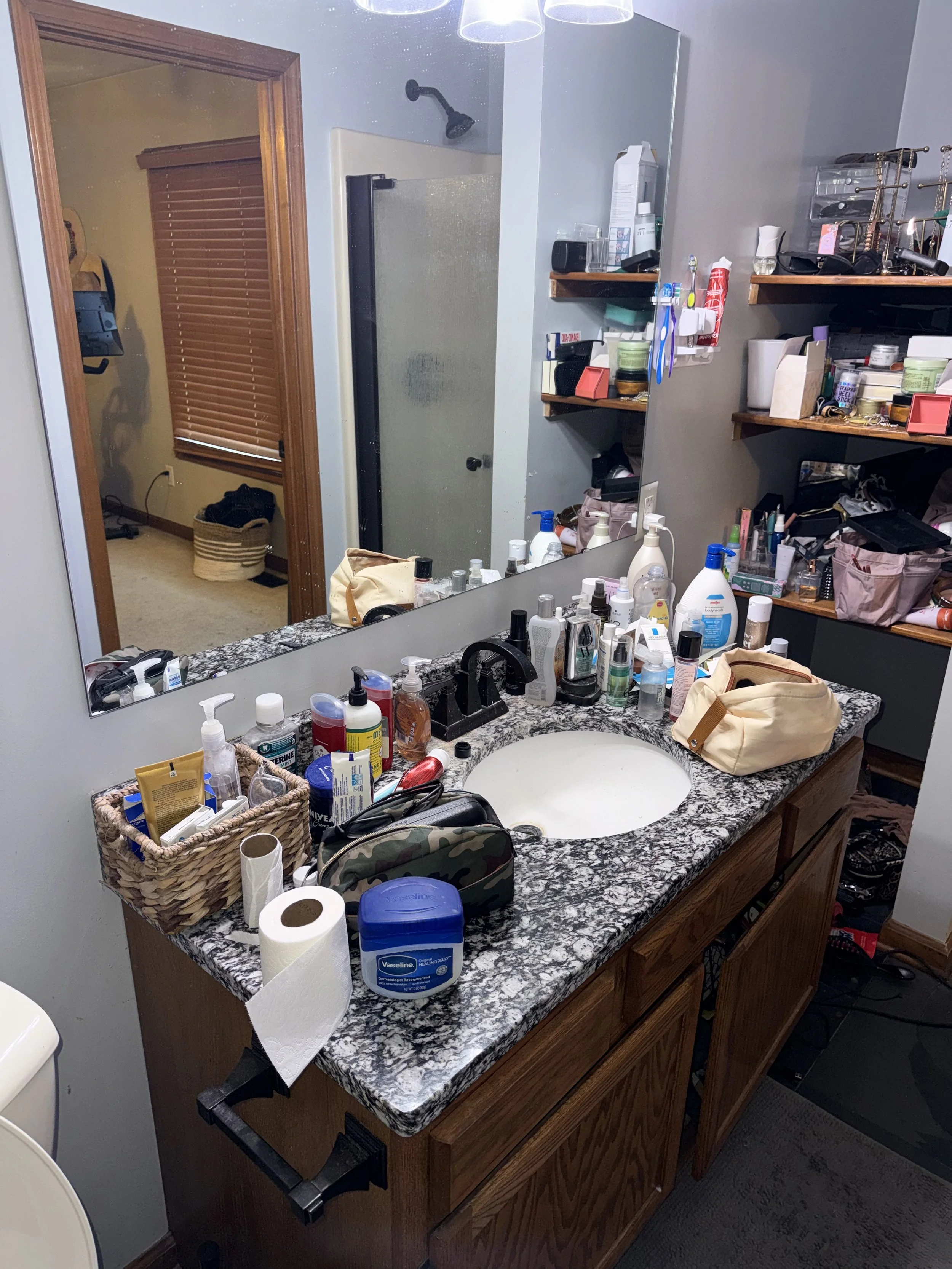 A cluttered bathroom countertop with various toiletries, a large mirror, and periodic shelves of personal items.