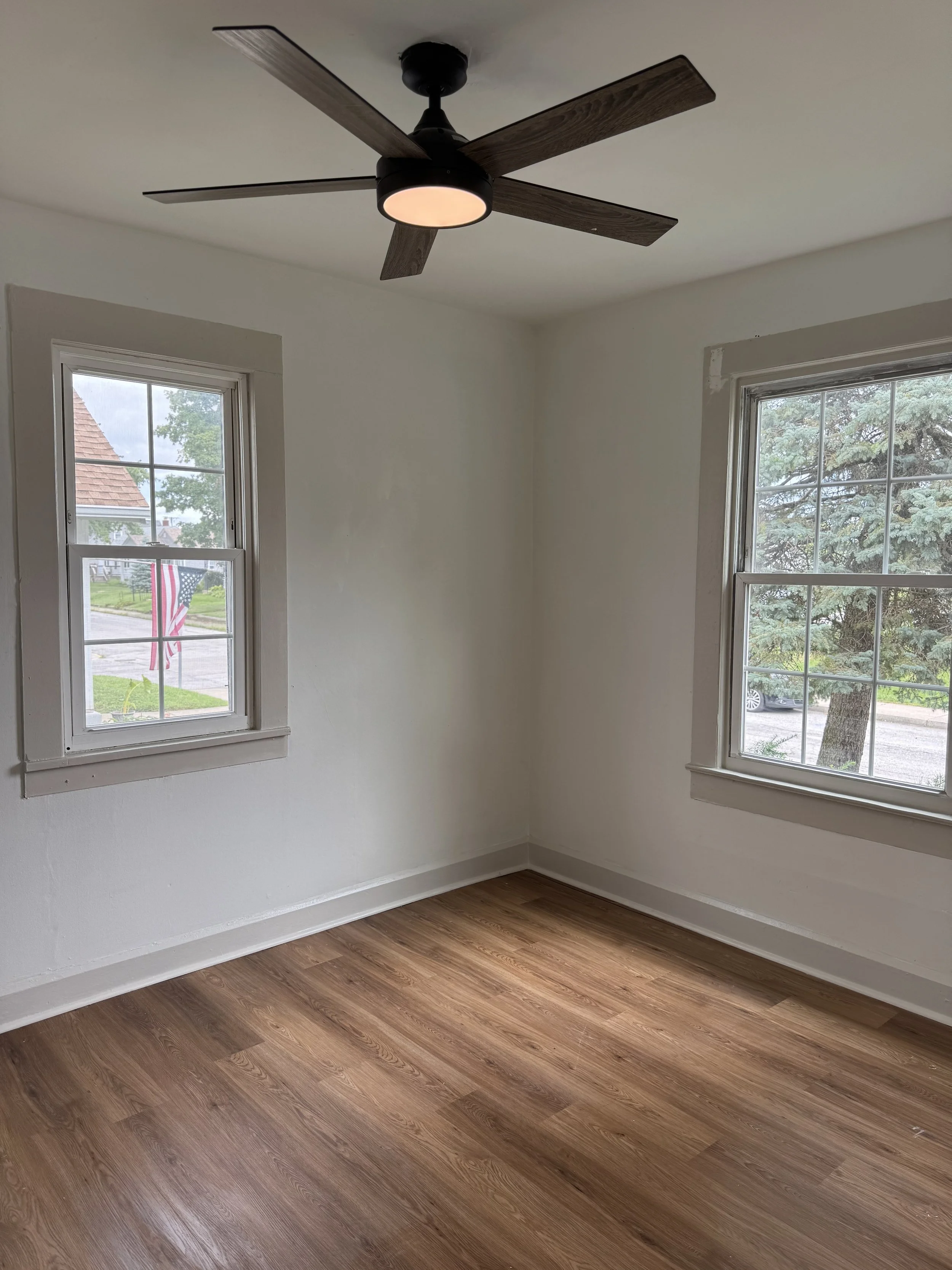 Empty room with white walls, two double-pane windows with white trim, a wood floor, a ceiling fan with dark wood blades and a built-in light, and a view of trees and neighboring houses outside.