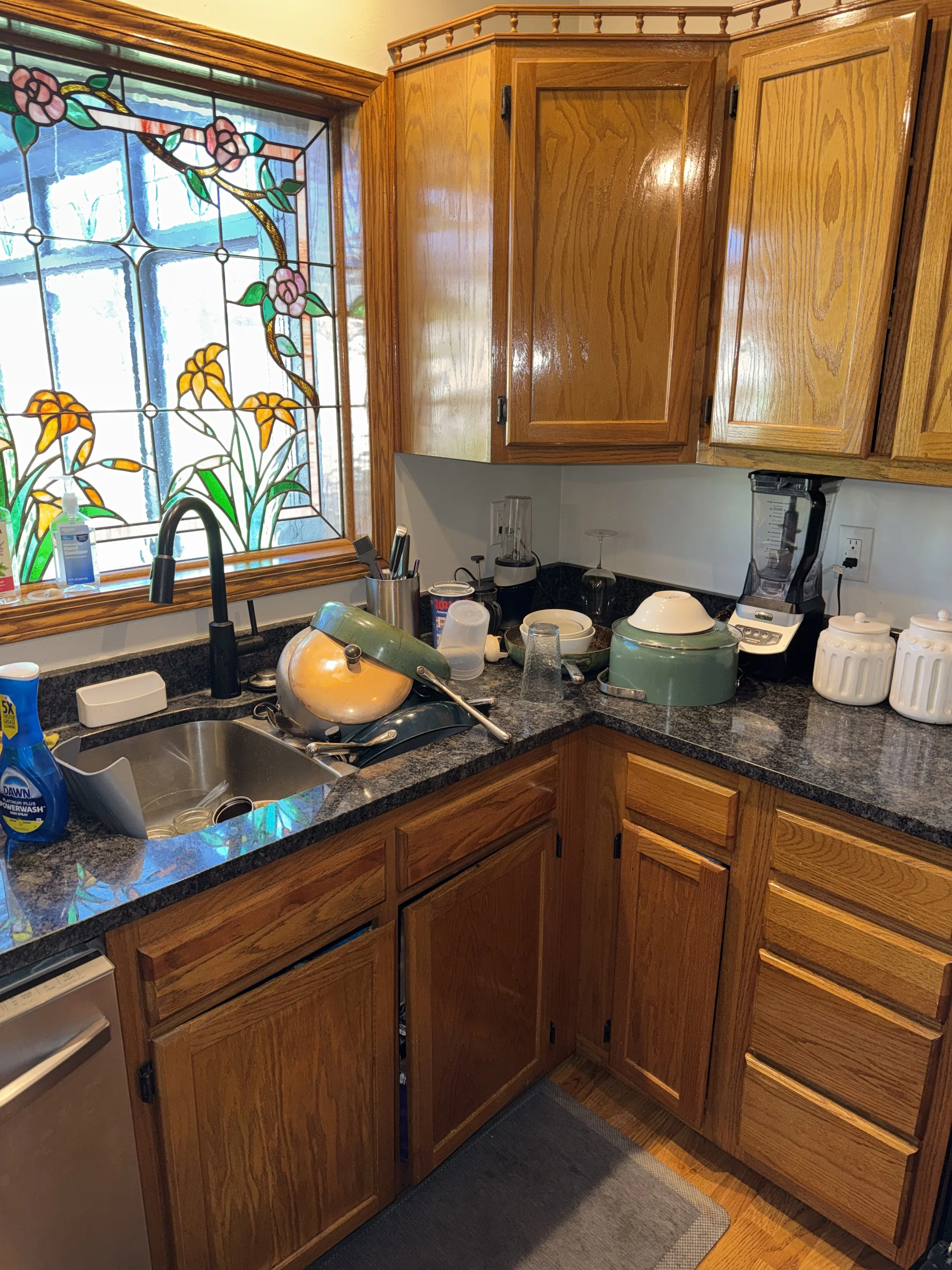 Cluttered kitchen counter with a sink, wooden cabinets, stained glass window, blender, and various dishes and containers.