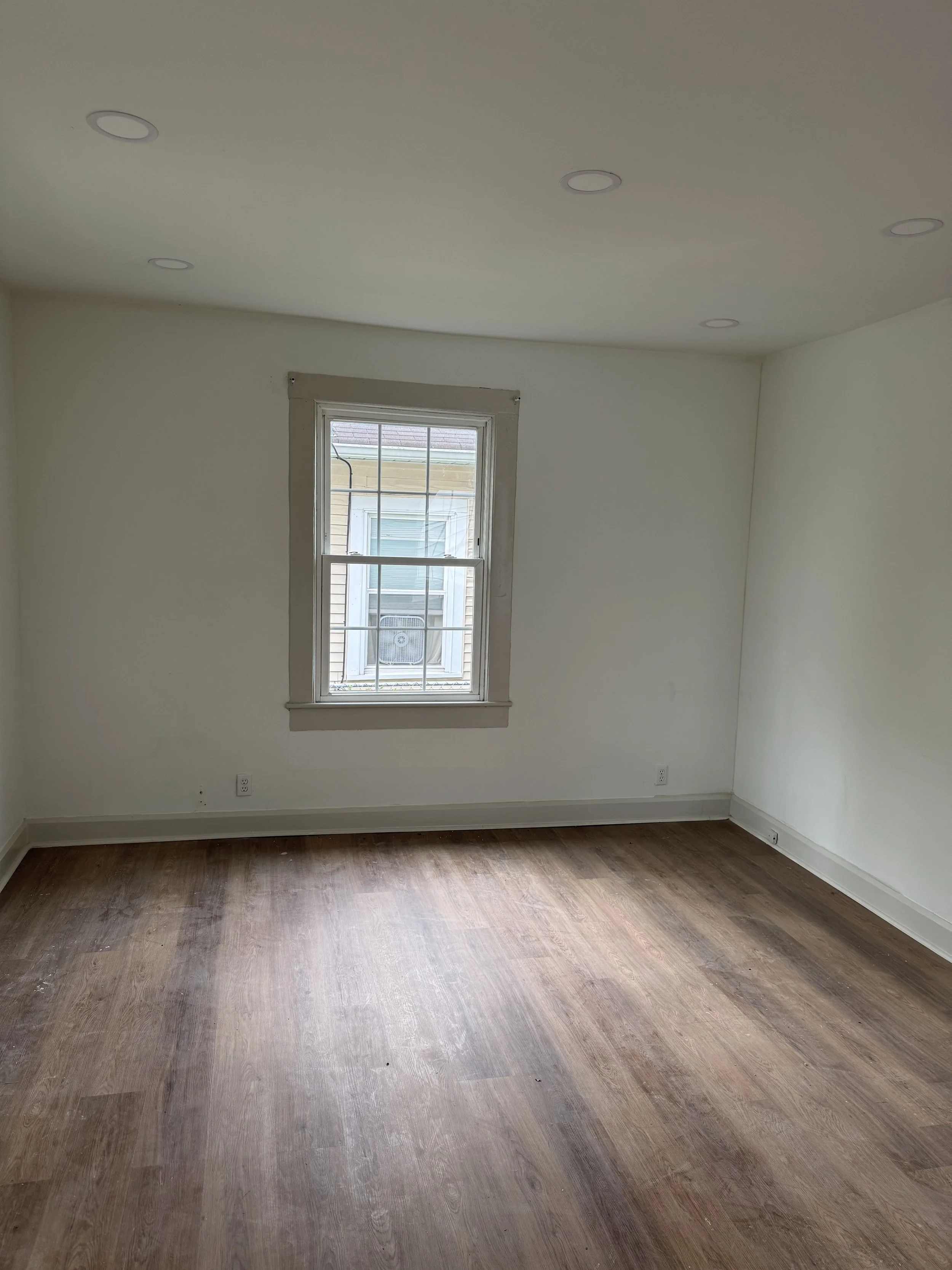 Empty room with wooden floors, white walls, and a single window with white frame and grid pattern.
