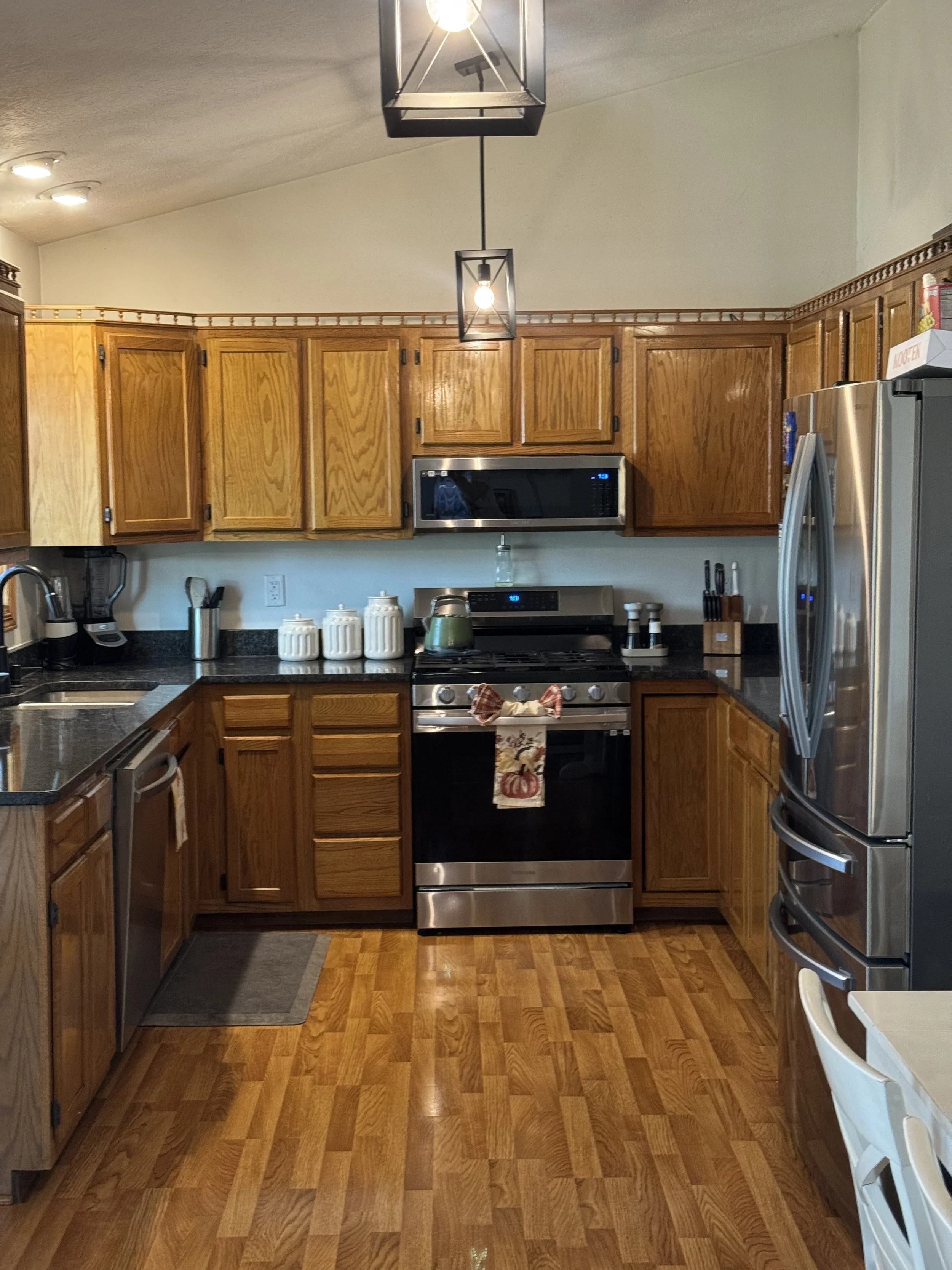 A kitchen with wooden cabinets, black countertops, stainless steel appliances including a refrigerator, microwave, and stove, and wooden flooring. There are various kitchen items on the counters, including jars, knives, and a coffee maker.
