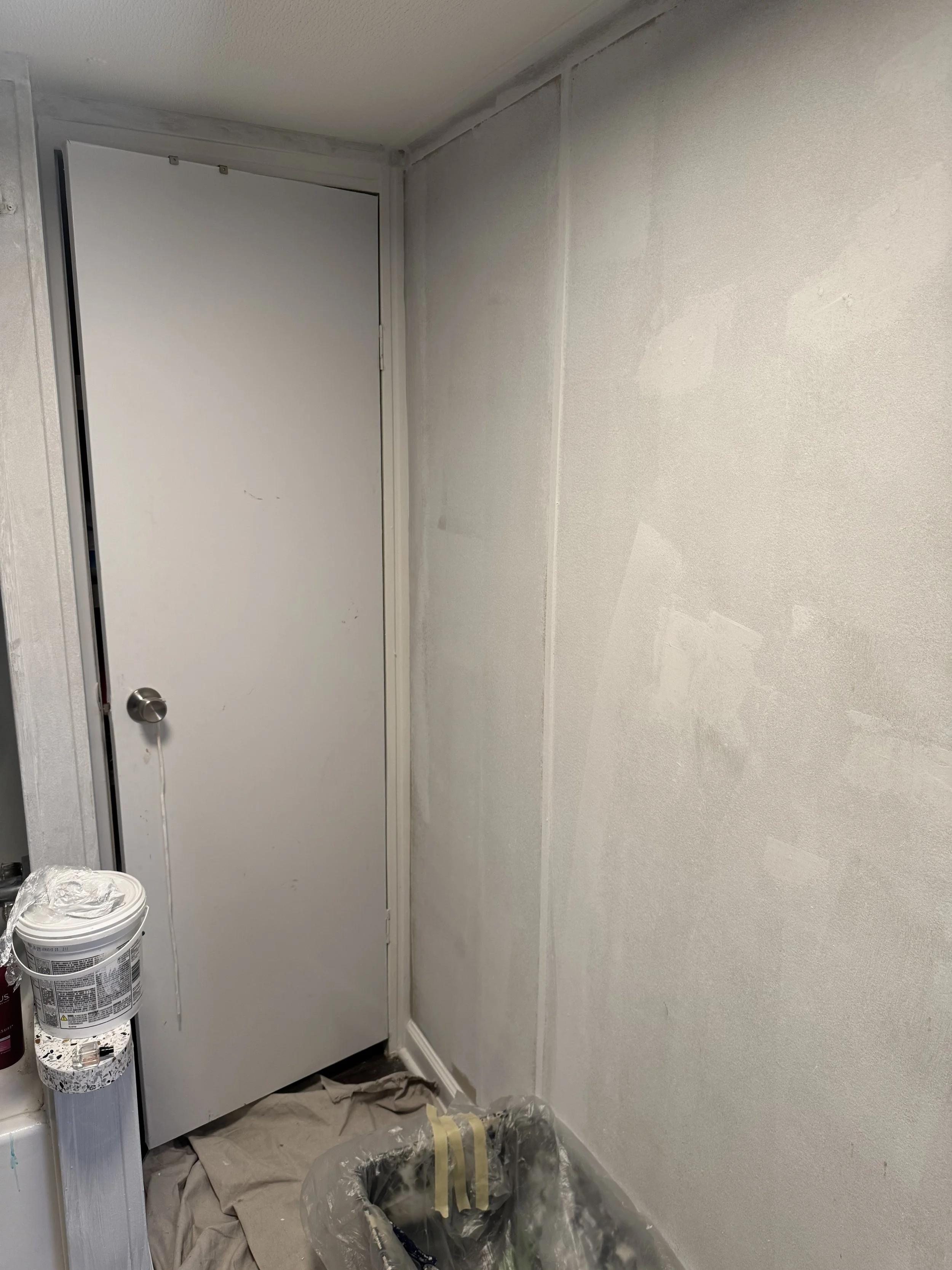 A white interior door with a round metal doorknob, surrounded by painted white walls with some patches of primer or paint, a bucket of paint, a plastic bag, and painter's tape on the floor.