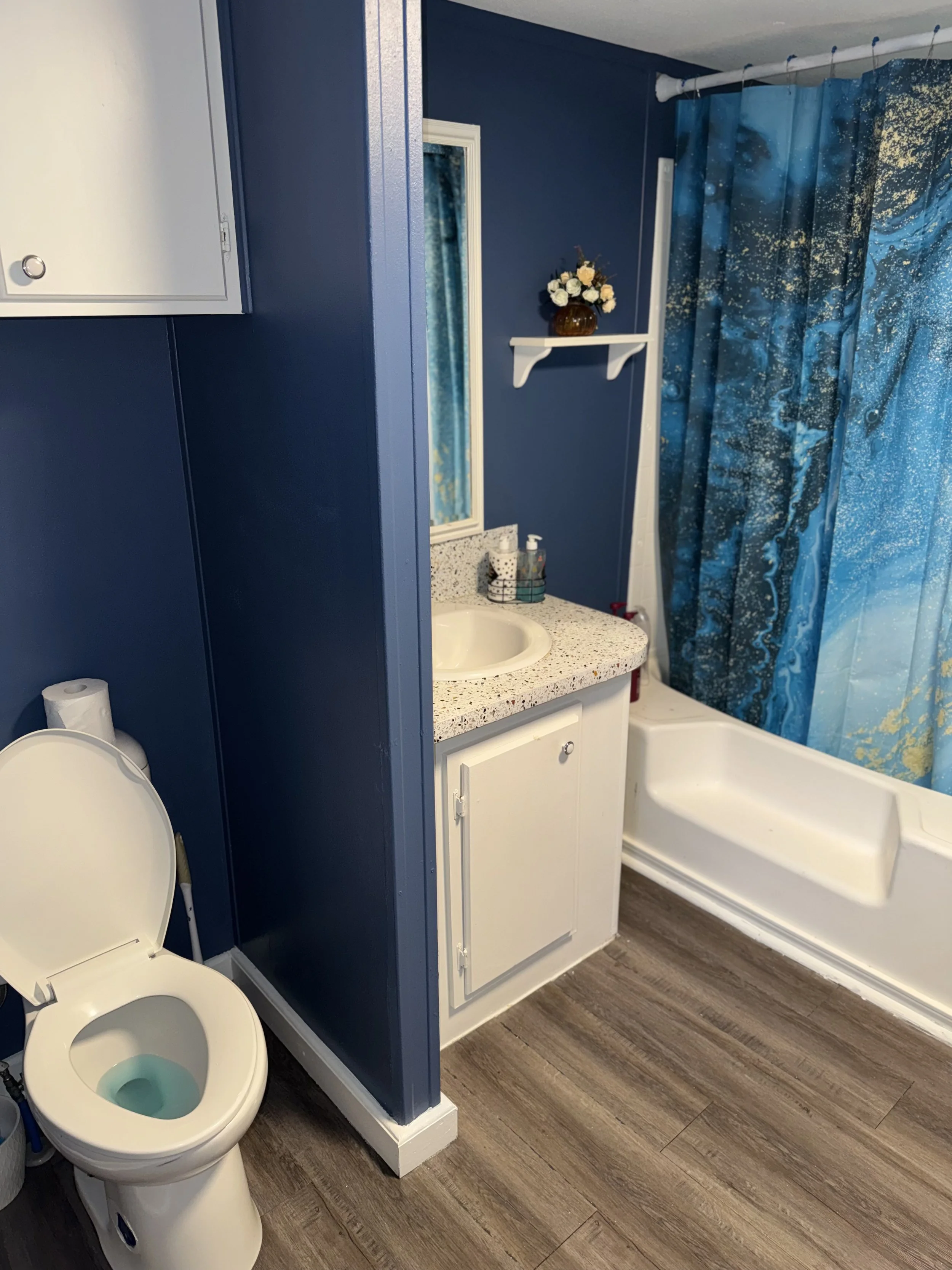 Bathroom with blue walls and a wooden floor, featuring a toilet with an open lid, a white vanity with a speckled countertop and a sink, a mirror, a floating shelf with a flower arrangement, and a bathtub with a blue and gold shower curtain.