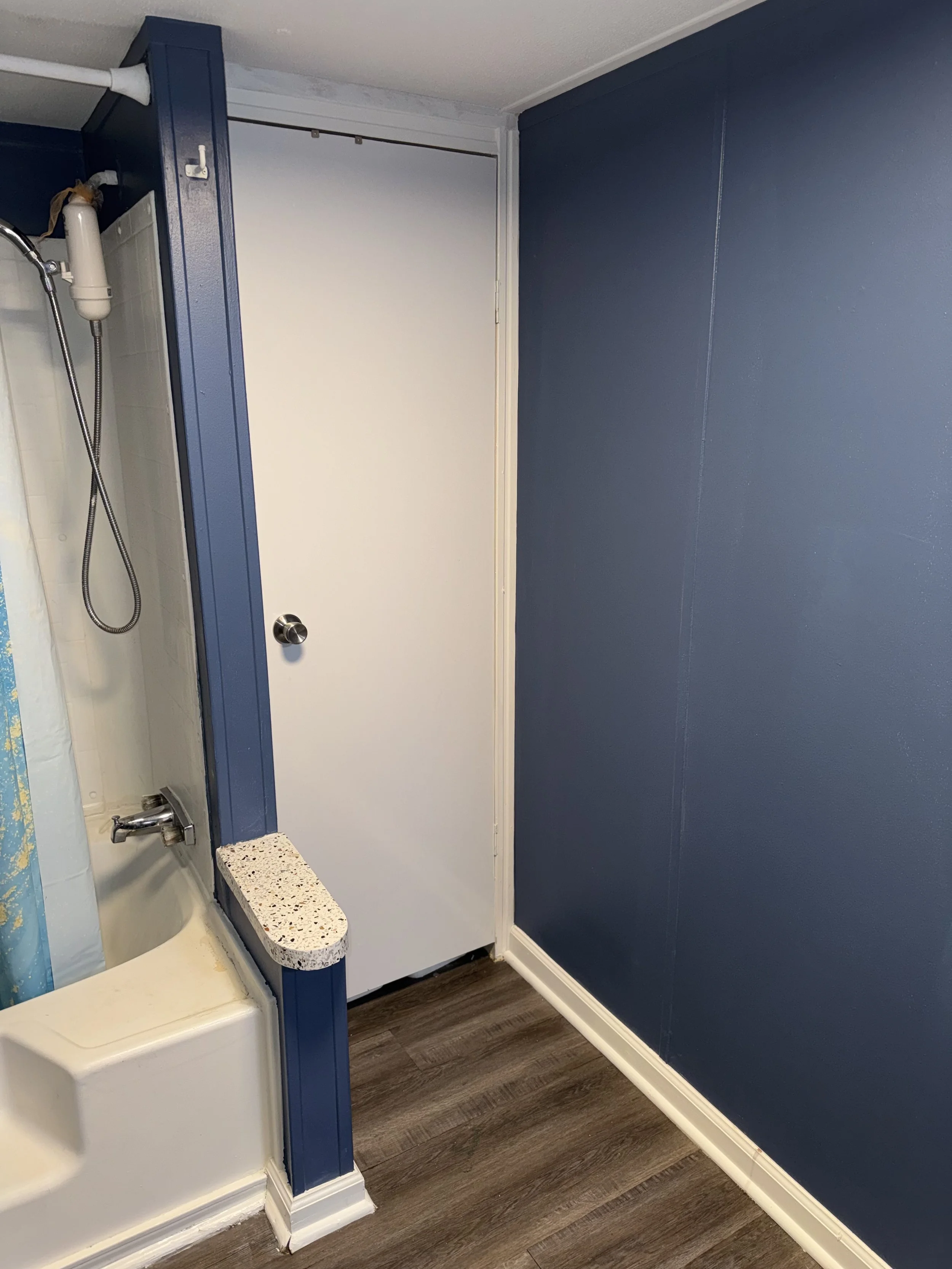 A corner of a bathroom showing a shower, a small shelf, a white door, blue walls, and wood flooring.