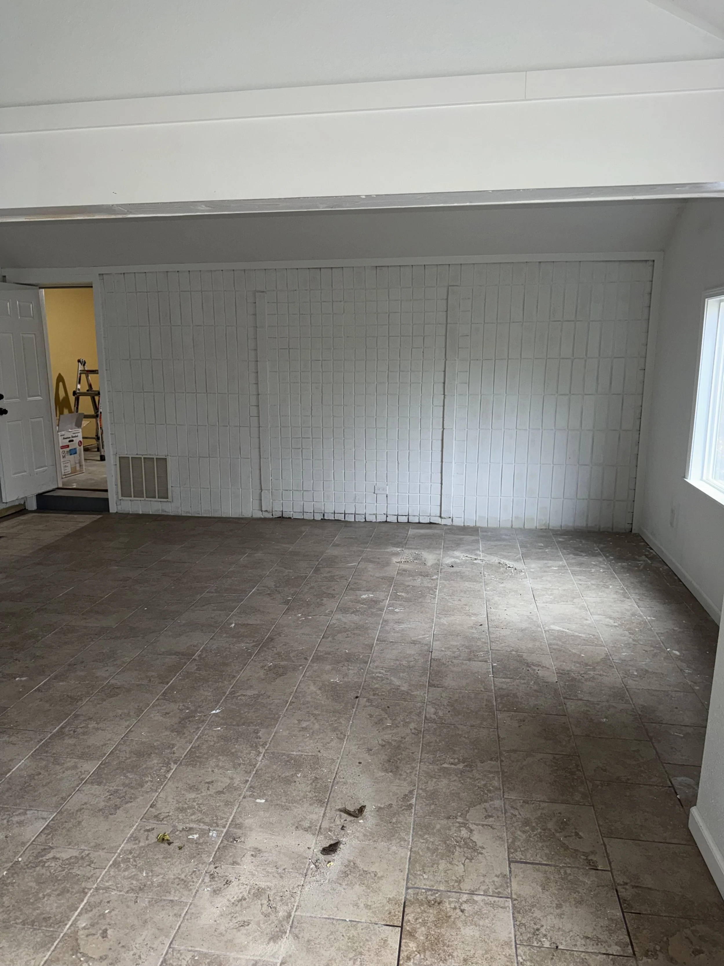 Empty room with tiled floor, white brick wall, window on the right, open door on the left revealing a small area with stairs and a ladder.