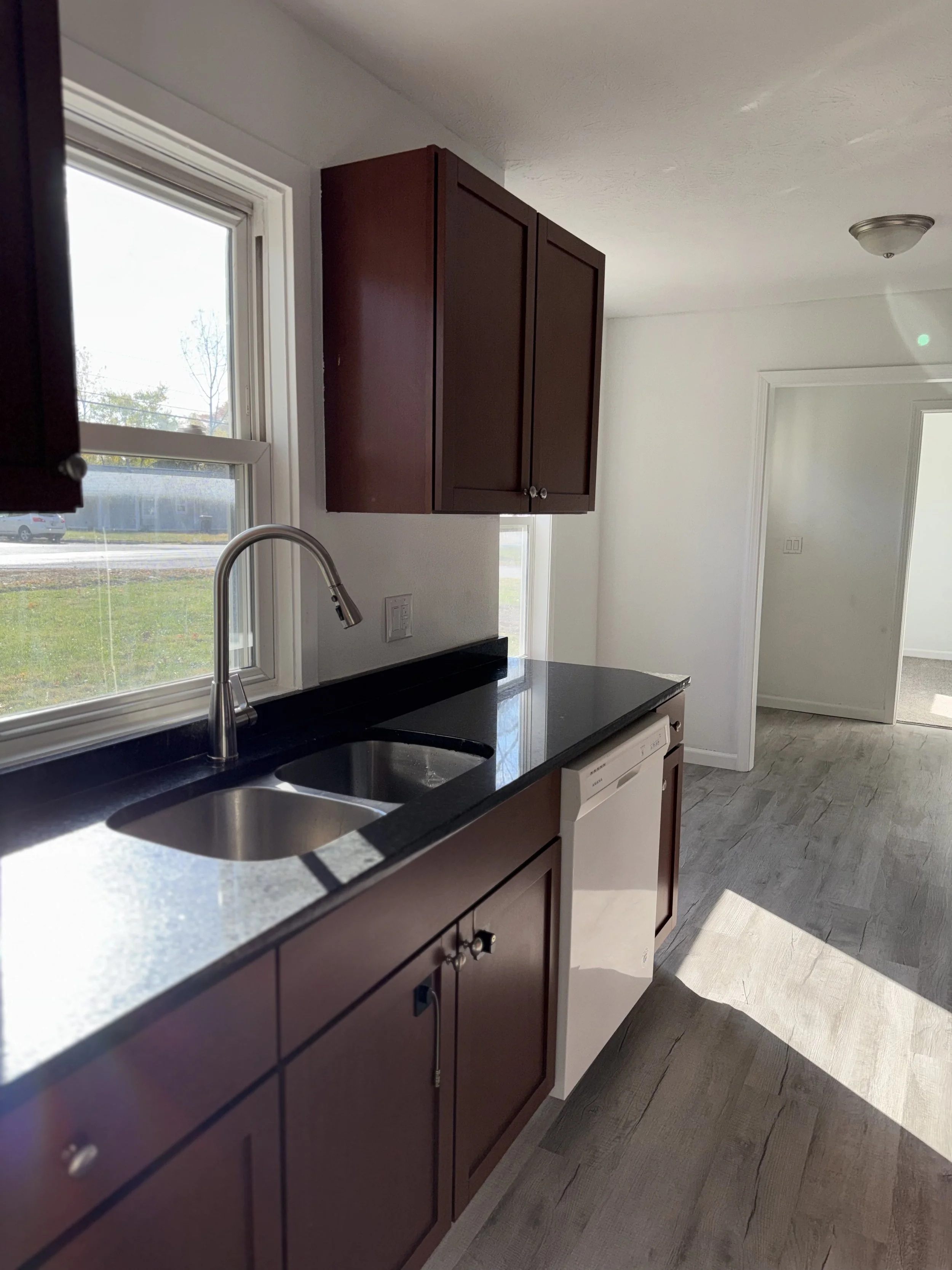 Kitchen with brown cabinets, black countertop, white dishwasher next to the sink, and a window above the sink letting in natural light. The floor appears to be wood or laminate.