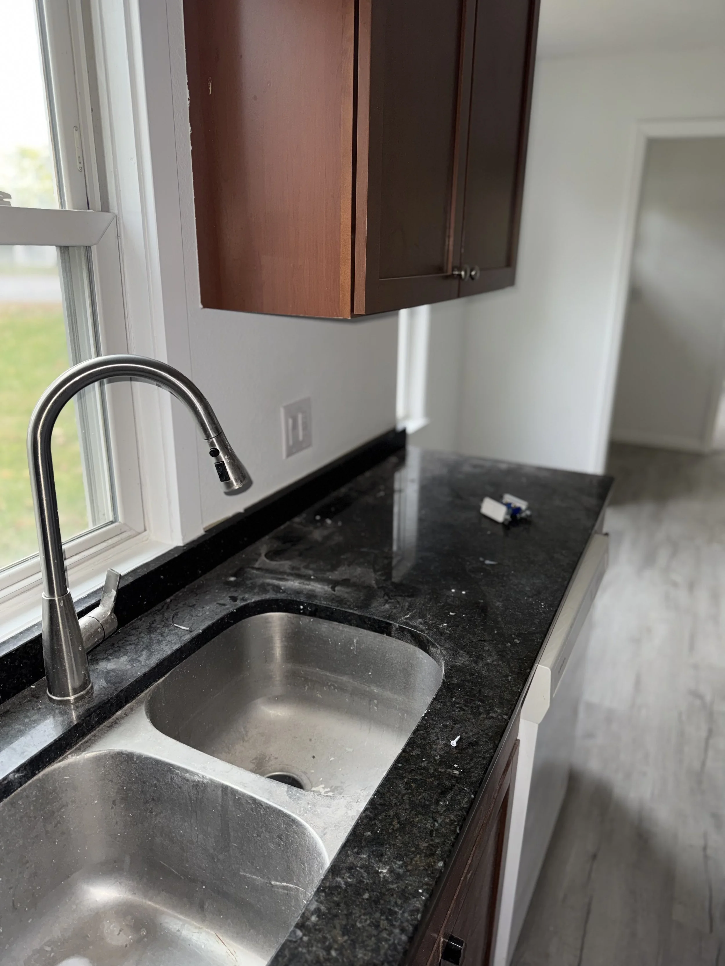 Kitchen with granite countertop, stainless steel sink, white walls, wood cabinets, and a window, with some debris on the counter.