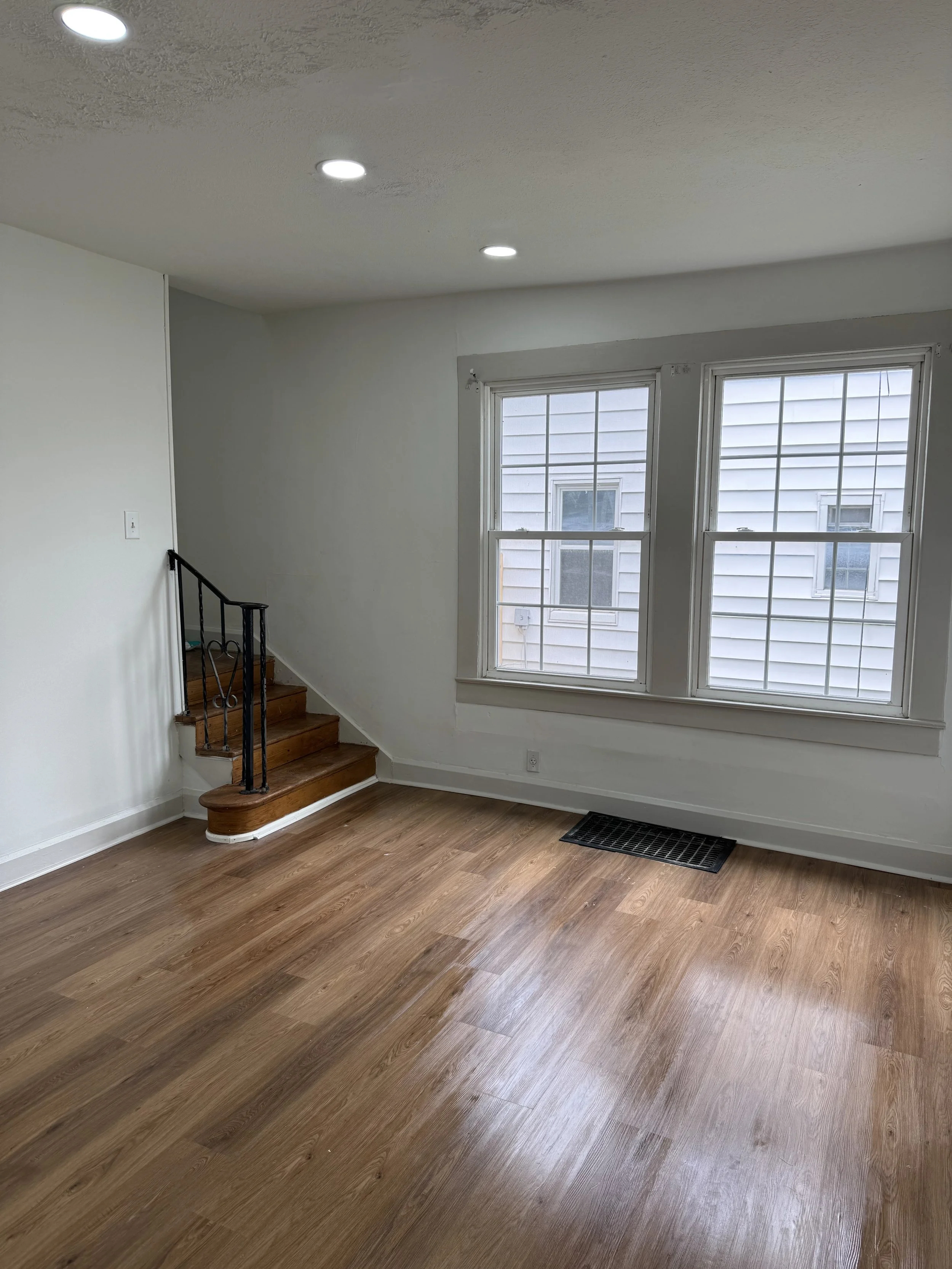 Empty living room with hardwood floors, white walls, two large windows, a small staircase with a black metal railing, and recessed ceiling lights.