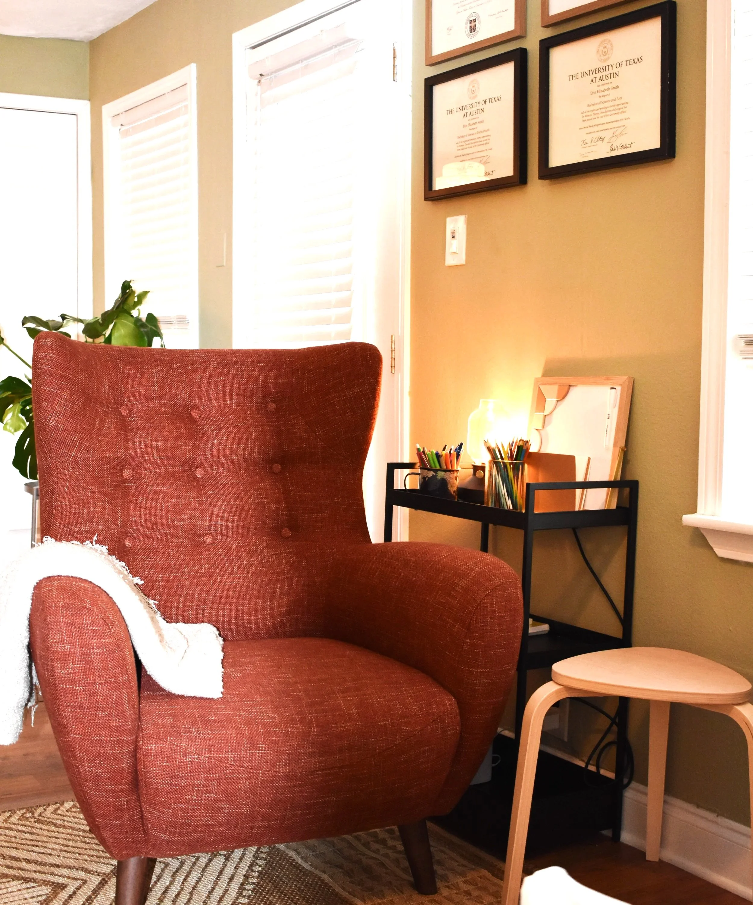 A cozy reading corner with an orange upholstered armchair, a small black side table with colored pencils, a lamp, and framed university diplomas on the wall.