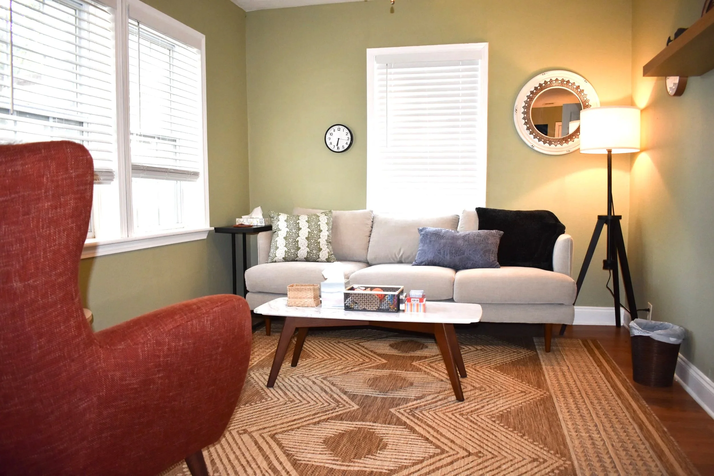 Living room with a beige sofa, dark cushions, floor lamp, a red armchair, a coffee table, window blinds, and wall decor.