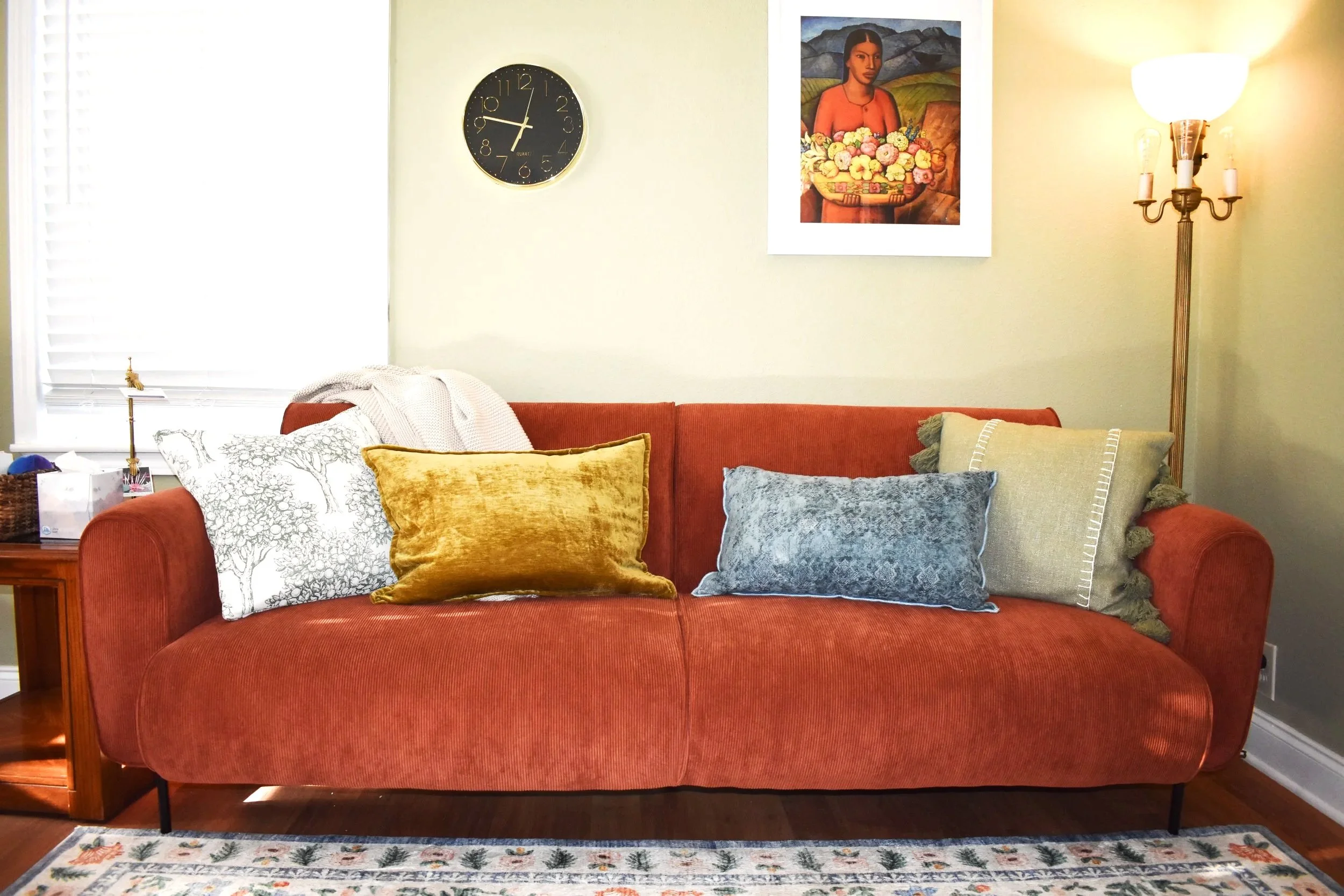 Living room with a coral sofa, various pillows, a wooden side table, a window with blinds, a black wall clock, framed artwork, and a standing lamp.