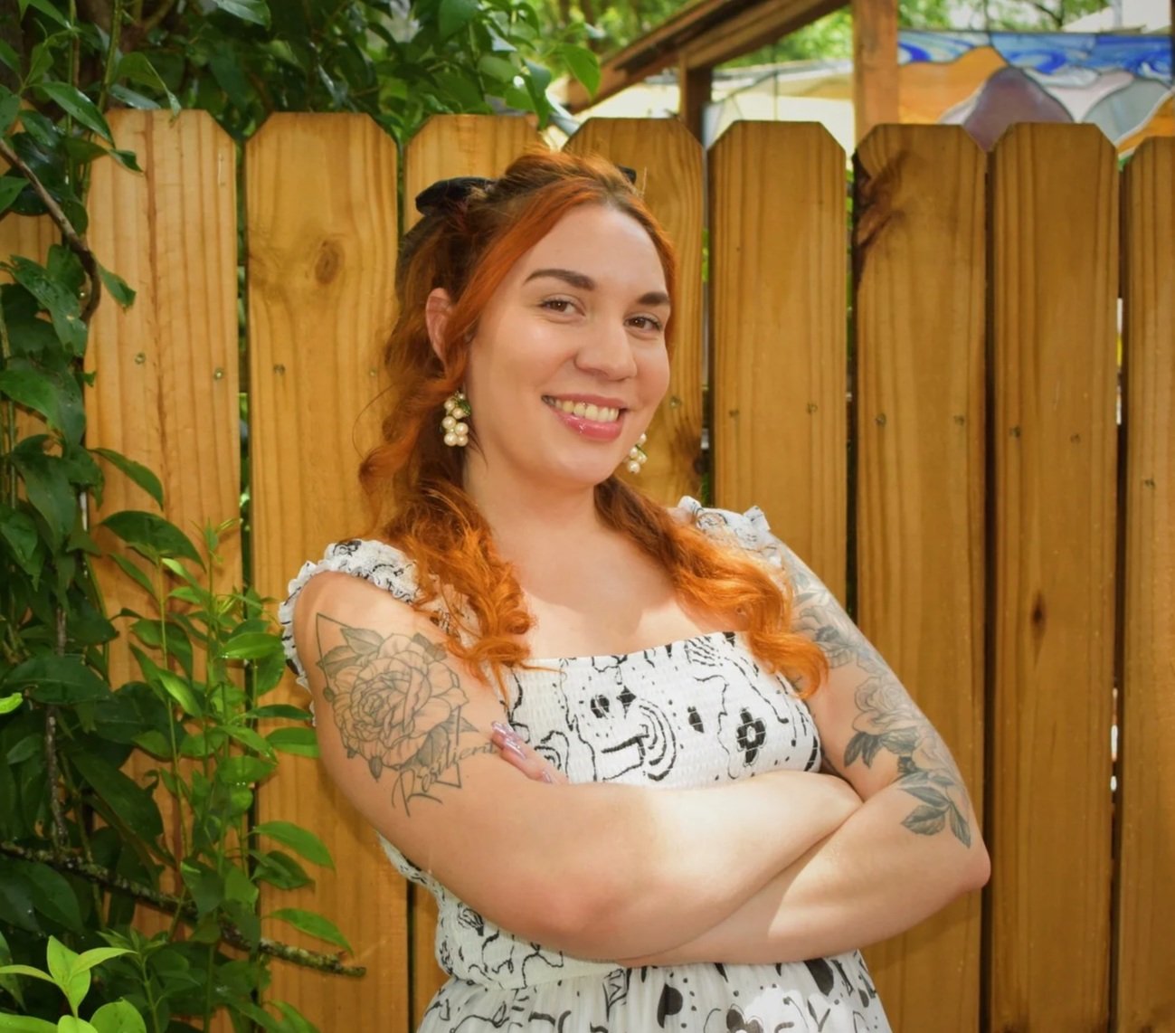 Marissa Sanchez, LCSW in Austin, TX. A woman with red hair and floral tattoos on her arms, smiling with arms crossed, standing outdoors in front of a wooden fence and greenery.