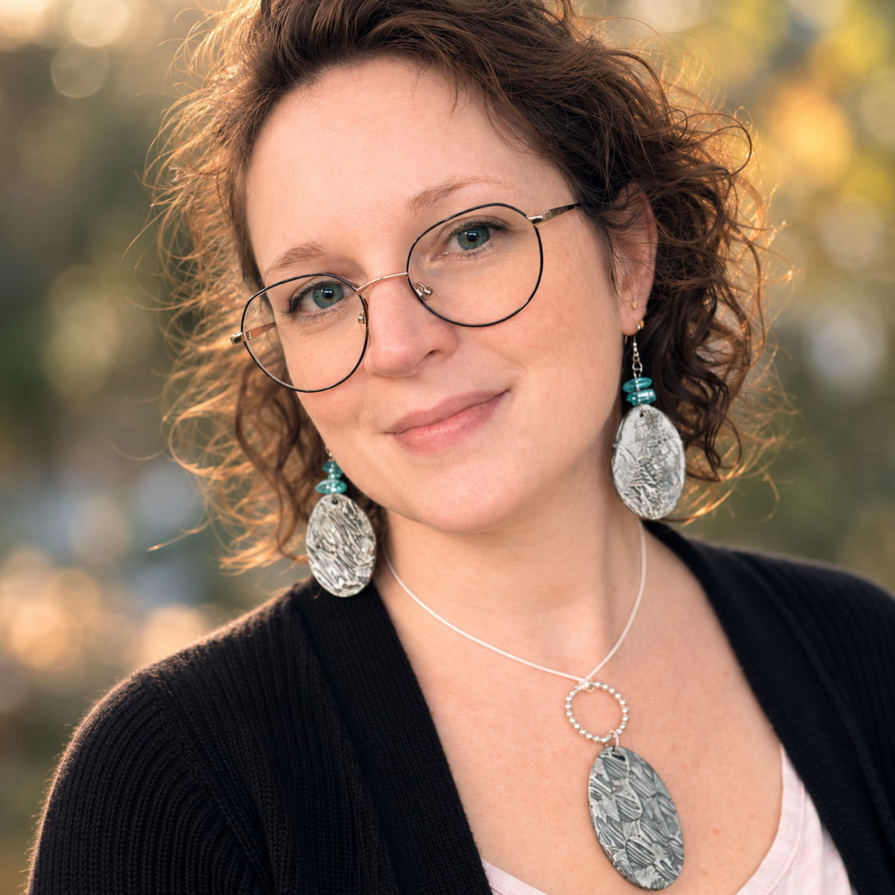 A woman with curly brown hair, glasses, and blue eyes wearing large silver and turquoise jewelry, smiling outdoors with blurred fall trees background.