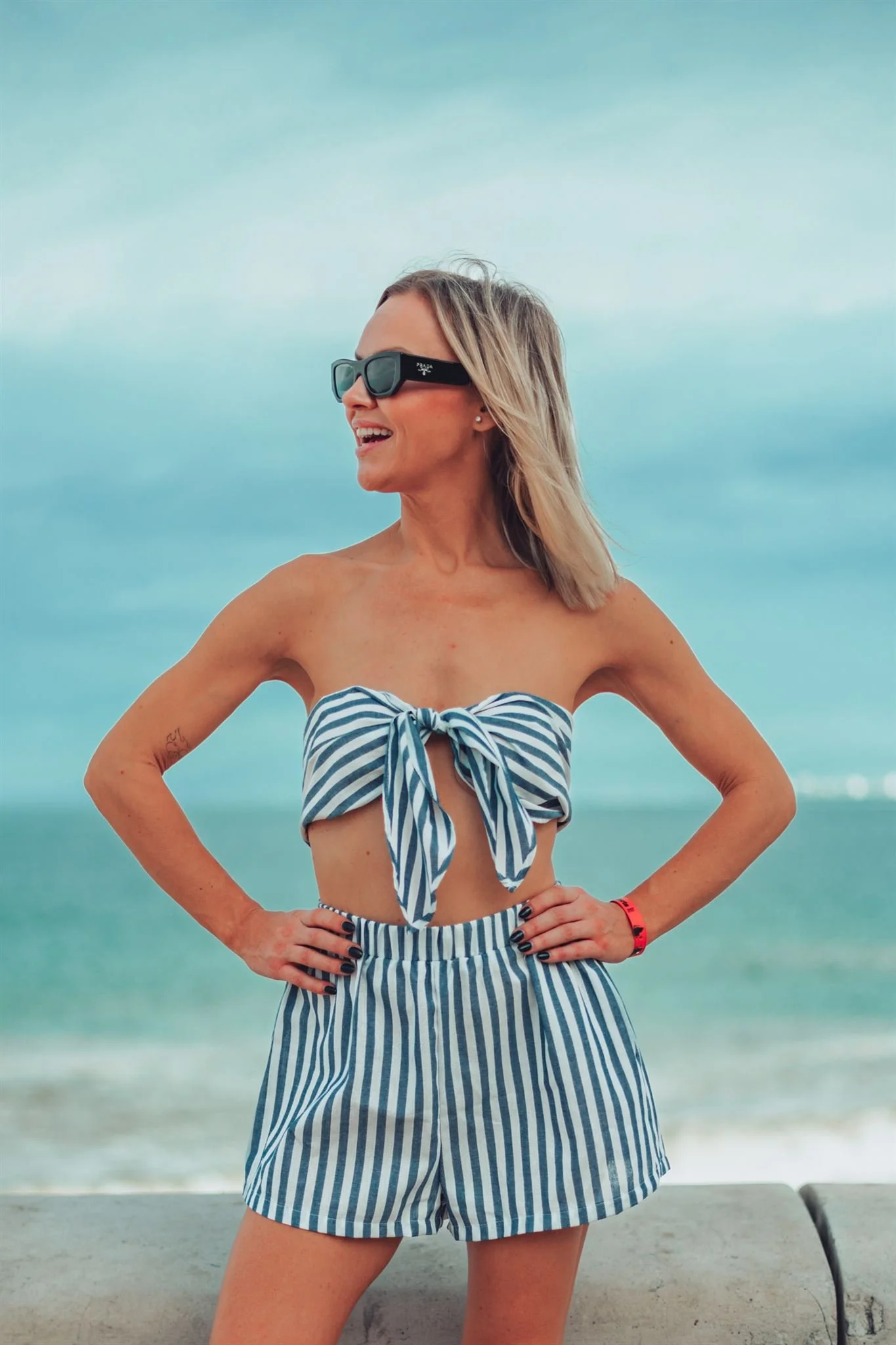 Nicole Shewaga smiling on a beach, wearing a striped blue and white outfit and sunglasses.