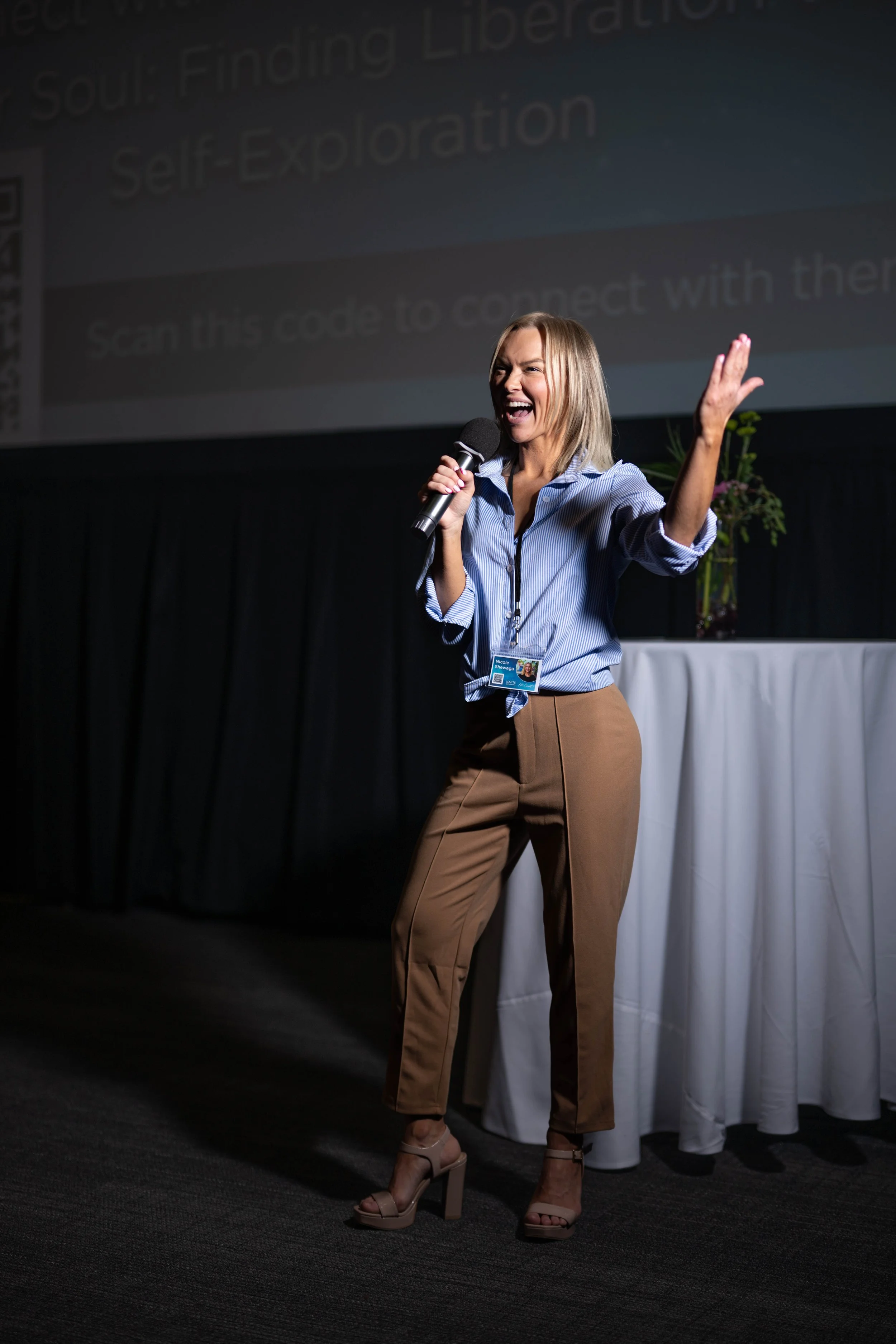 Nicole Shewaga speaking on stage with a microphone, smiling and gesturing toward the audience.