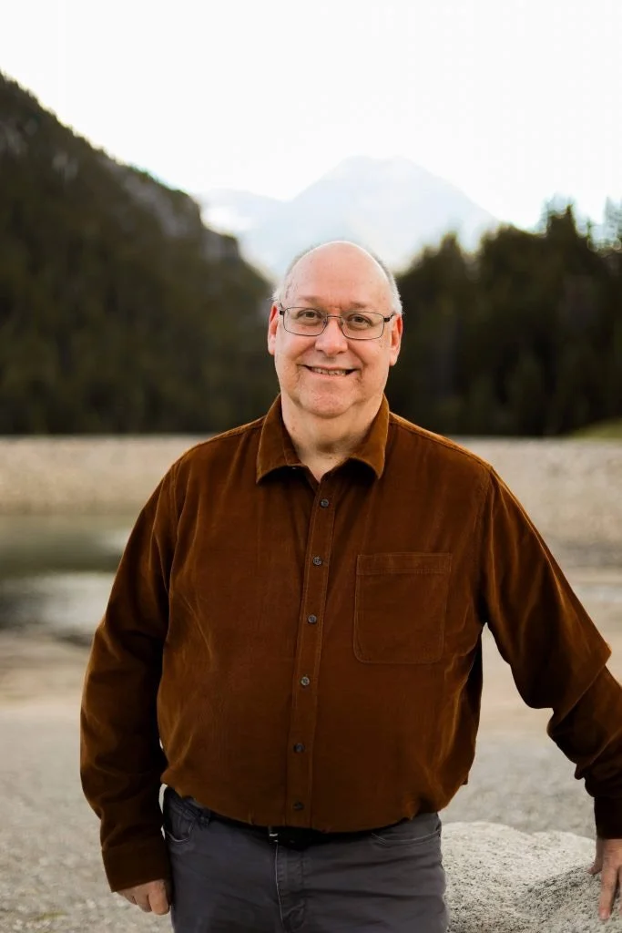A smiling man with glasses and a brown shirt standing outdoors near a river with mountains in the background.