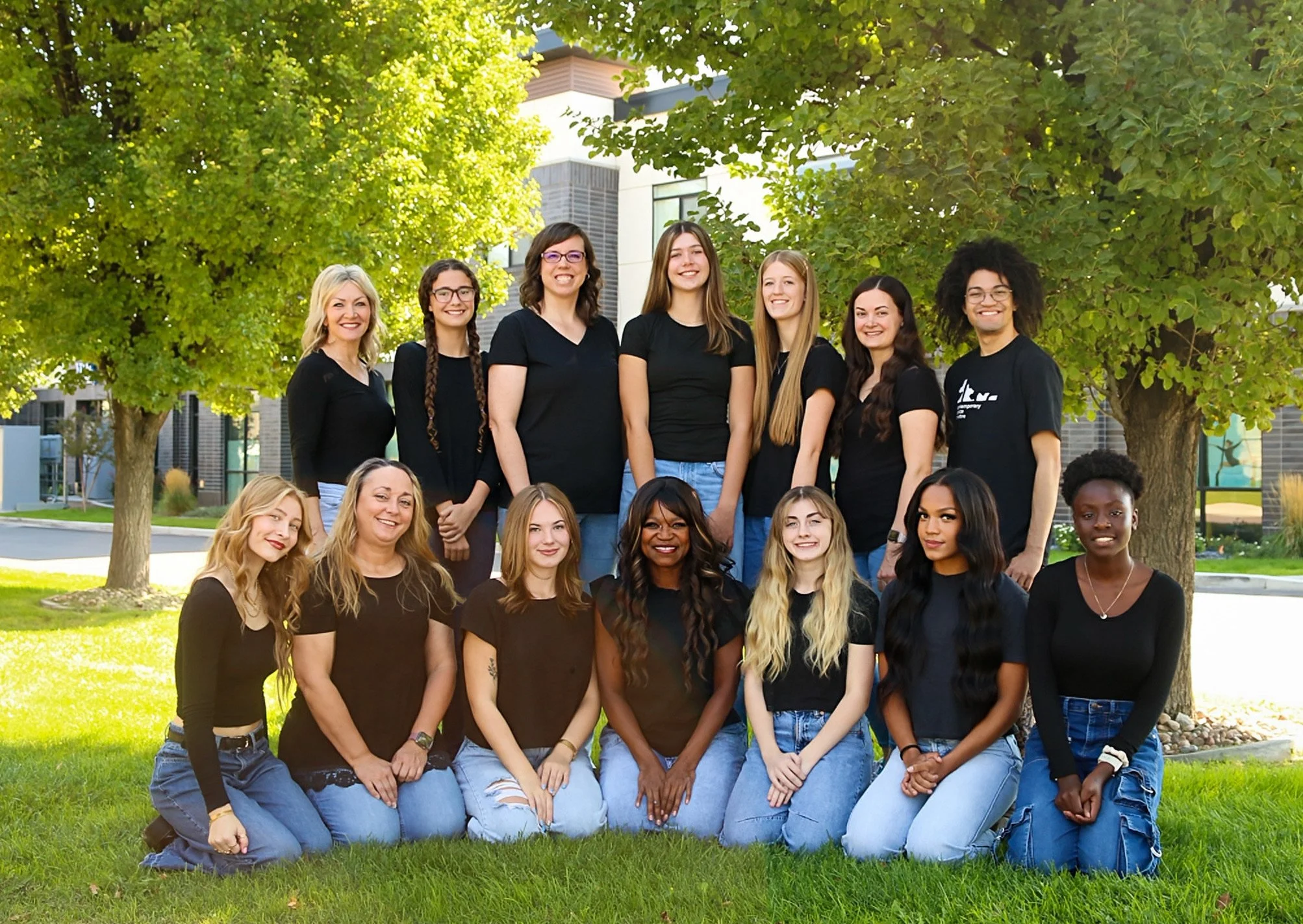 A diverse group of fifteen women posing outdoors in front of trees and a modern building, all wearing black shirts and jeans.