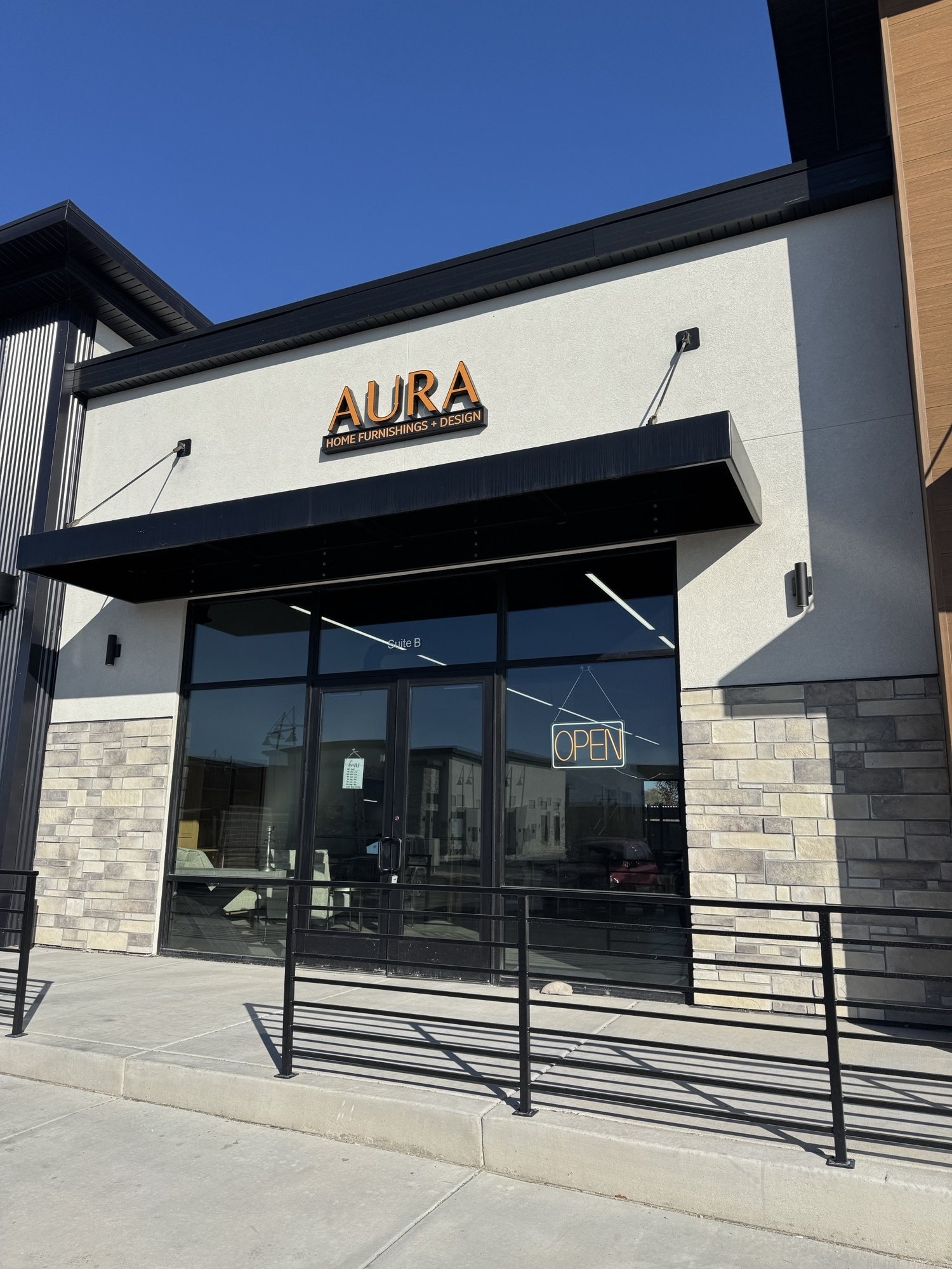 Exterior of a furniture store called Aura, with a sign that reads 'HOME FURNISHINGS + DESIGN'. The store has large glass windows, an 'OPEN' sign hanging inside, and black metal railings outside. The sky is clear and blue.