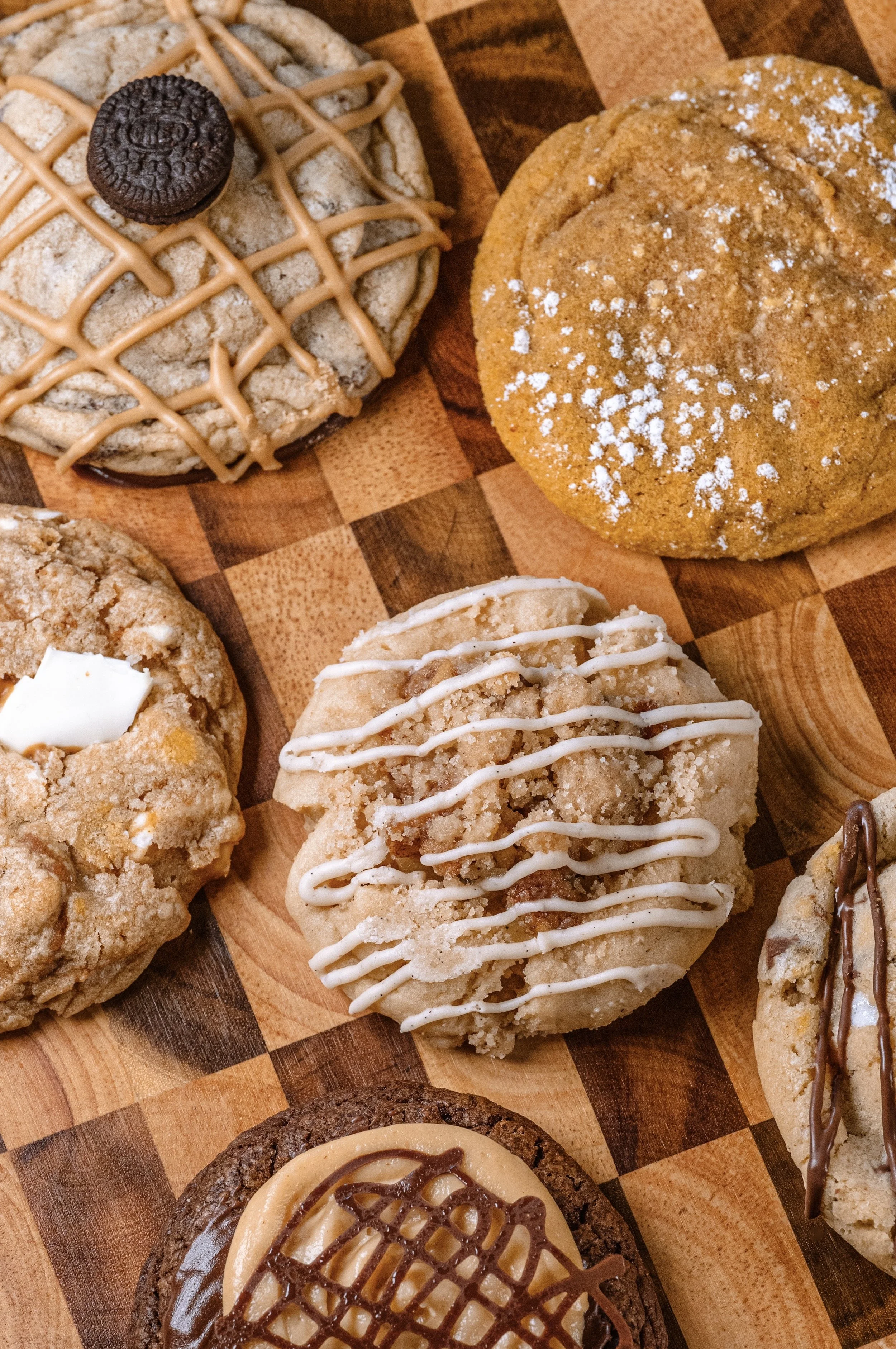 Assorted cookies on a checkered wooden surface, including an Oreo-topped cookie, a powdered sugar sprinkled cookie, a white drizzle decorated cookie, and a caramel-topped cookie.