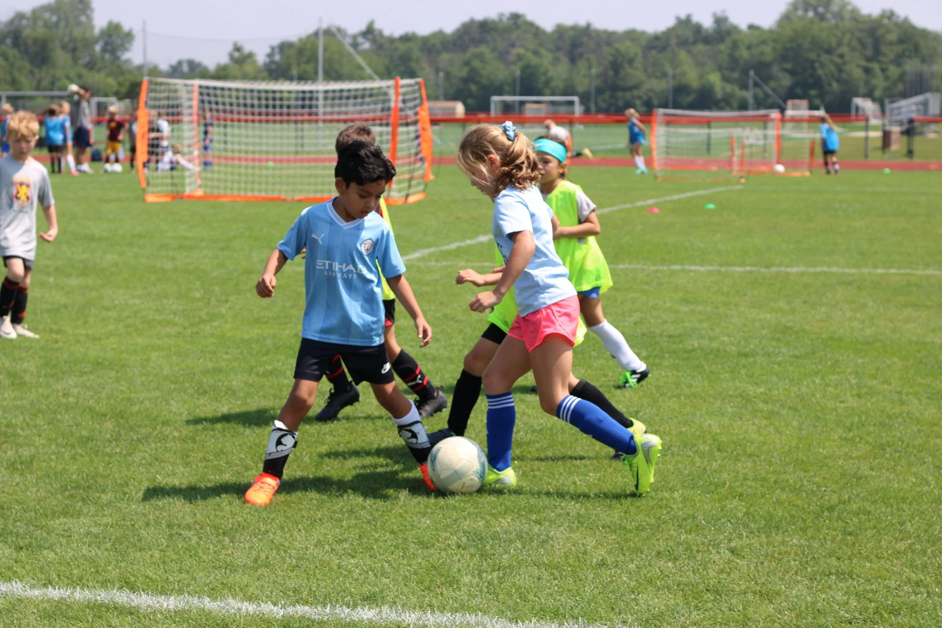 Children playing soccer on a grassy field during a sunny day, with soccer goals and other kids in the background.
