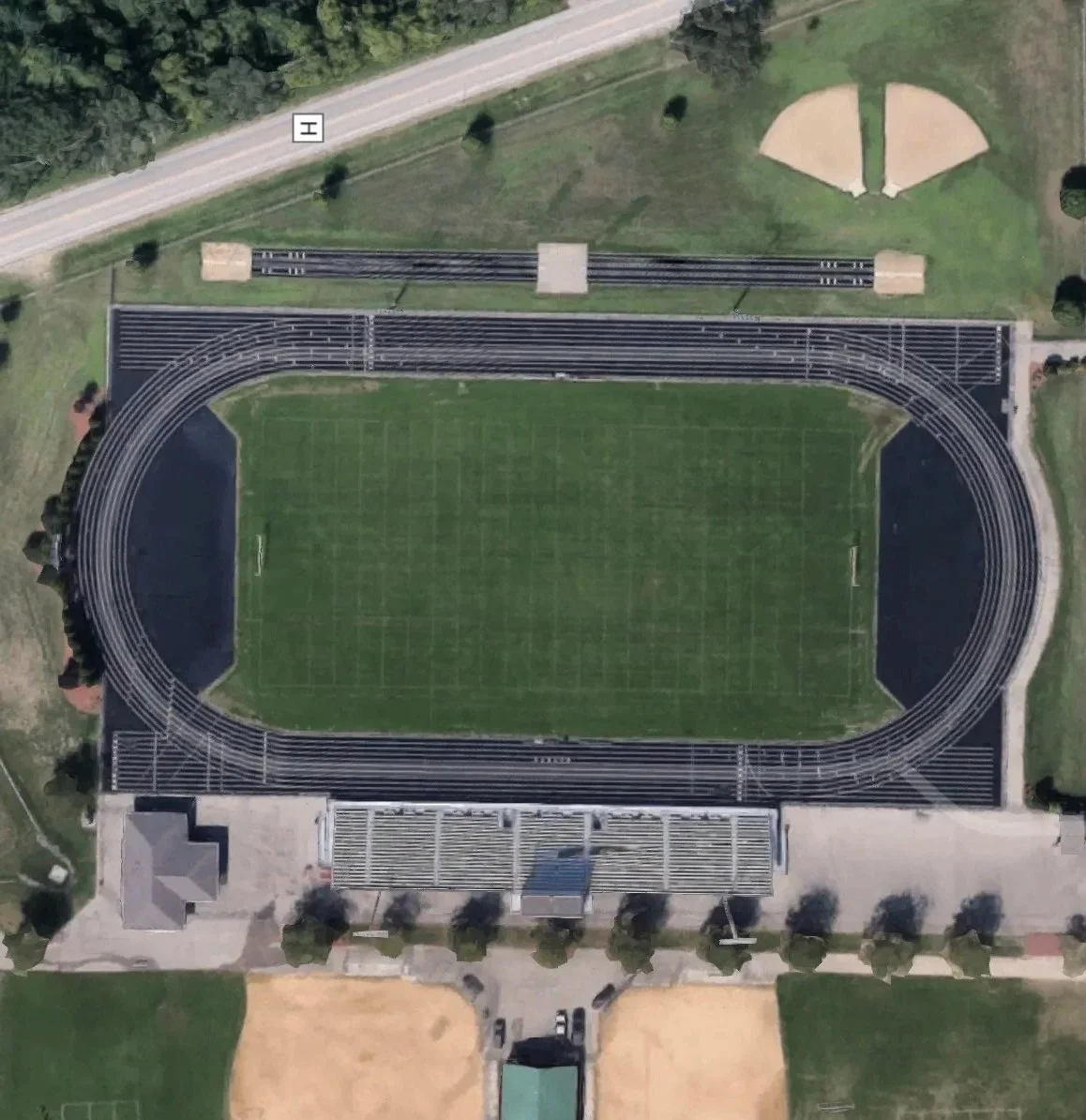 Aerial view of a sports stadium with a green field, surrounded by a running track with multiple lanes, bleachers on one side, and playgrounds and trees nearby.