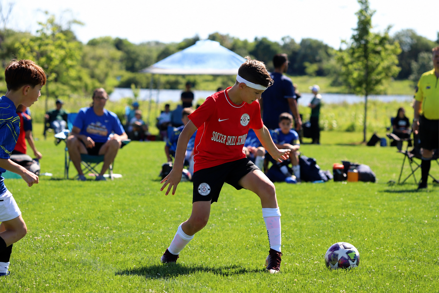 Young boys playing soccer on a field, wearing red jerseys with 'ILLINOIS SKILL SCHOOL' written on them, with a soccer ball on the grass, and spectators in the background. 3v3 academy. Soccer Skills Schools