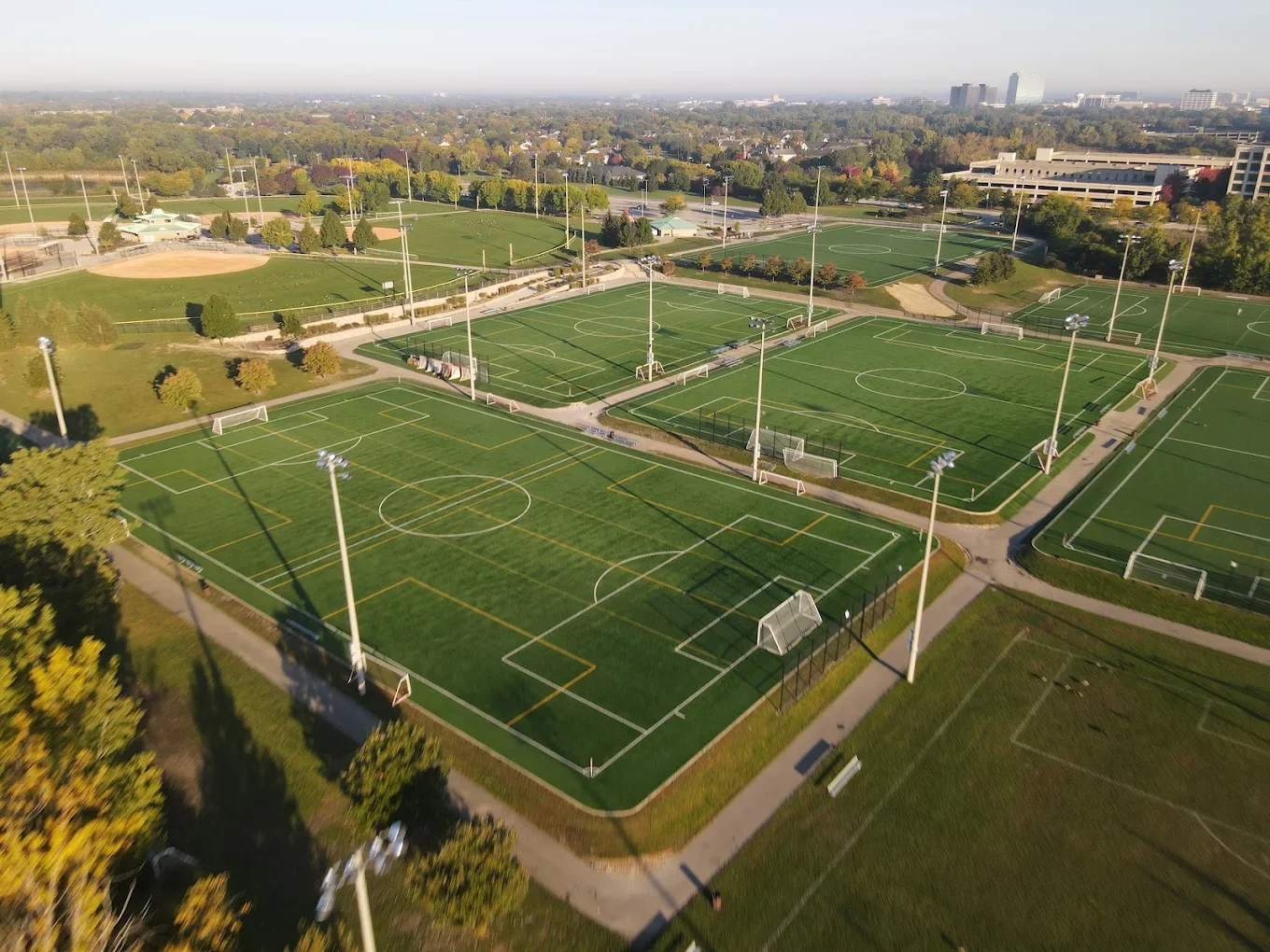 Aerial view of multiple sports fields, including soccer and football, illuminated by tall stadium lights, with a park, trees, and cityscape in the background on a clear day.