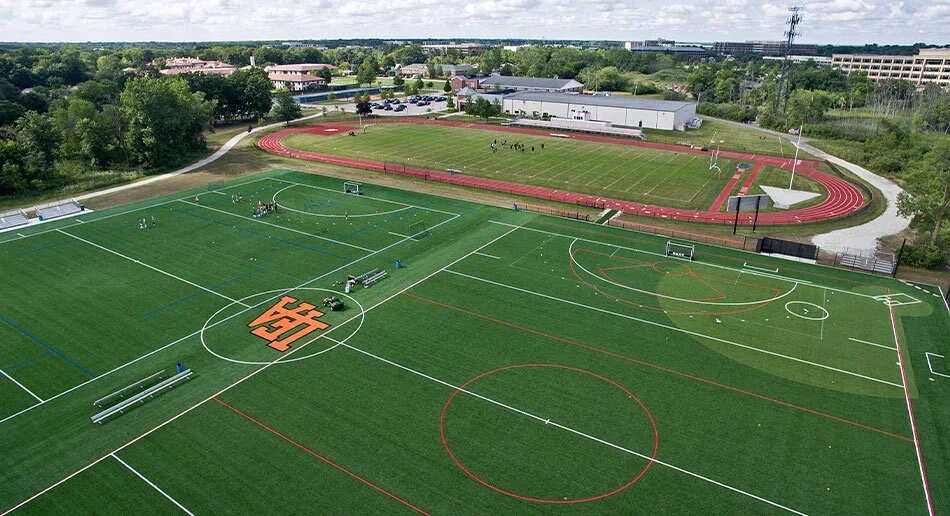 Aerial view of a sports complex with soccer fields, a running track, and a smaller basketball court, surrounded by trees and buildings.