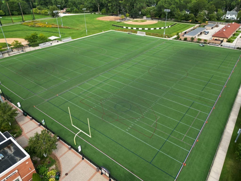 Aerial view of a large sports field with football and soccer markings, baseball field, and surrounding green space, in a park-like setting.