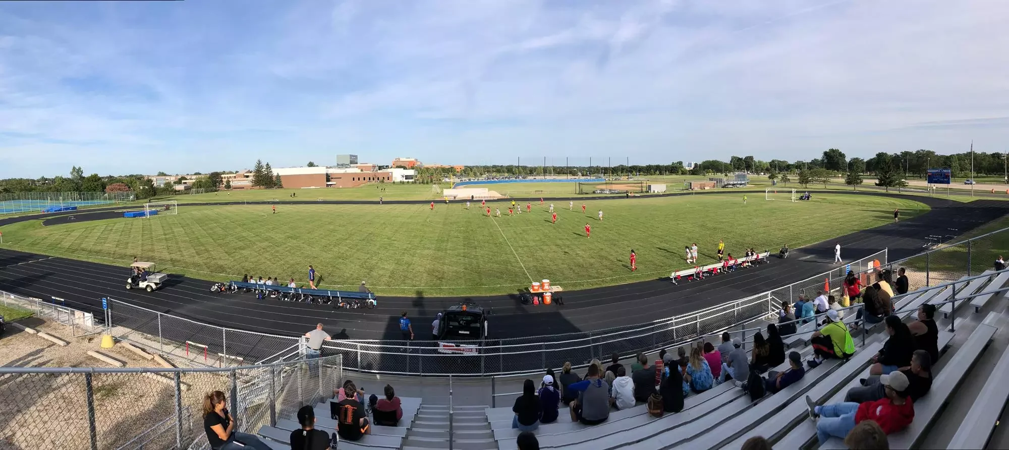 Panoramic view of a soccer field with players in red and white jerseys, spectators in the bleachers, and a track surrounding the field on a clear day.
