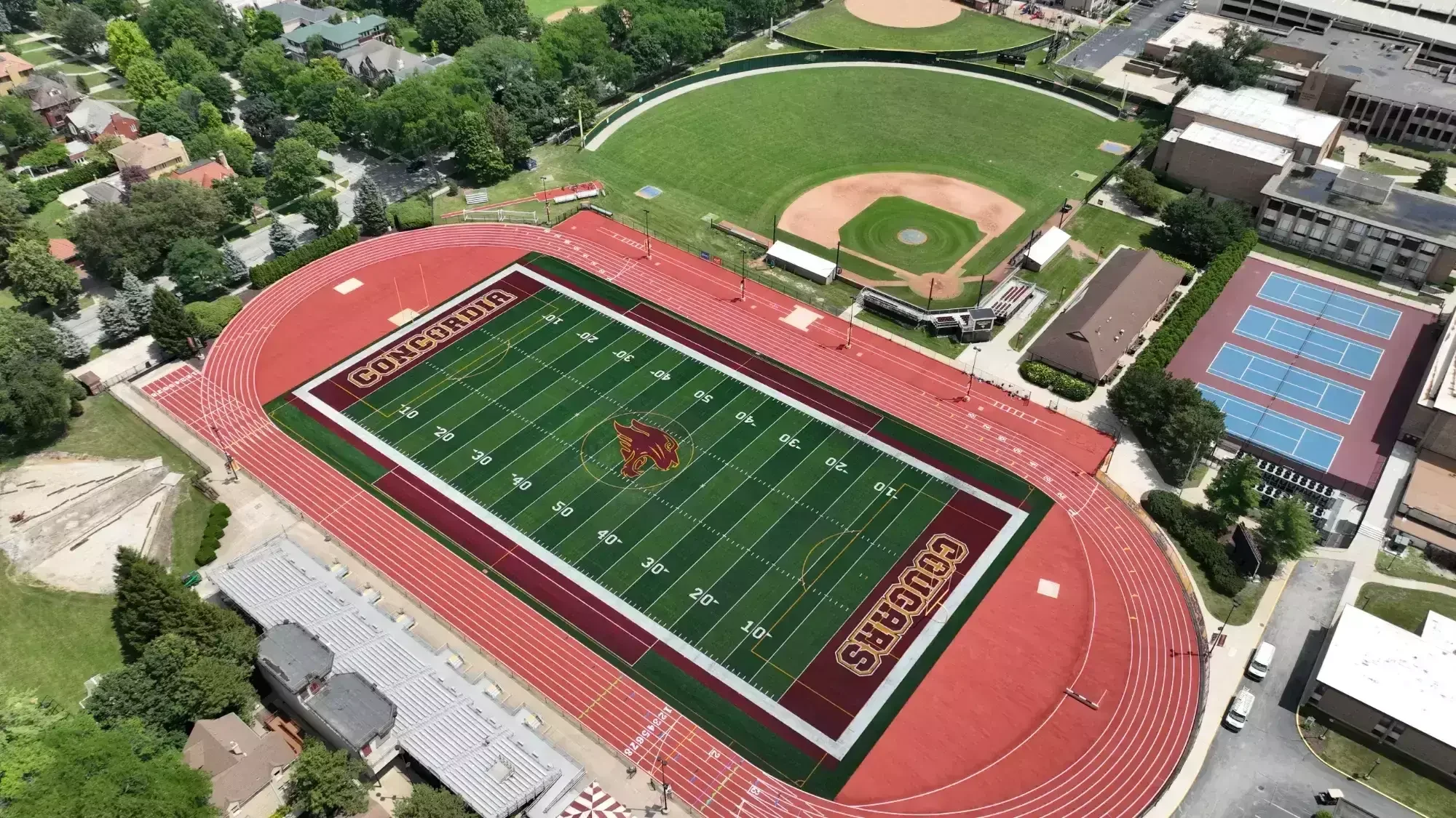 An aerial view of a sports complex with a football field in the center, surrounded by a running track. The field has the word 'CONCORDIA' and a mascot logo in the end zones. Adjacent to the football field is a baseball diamond, and nearby are tennis courts and a parking area with vehicles and buildings surrounding the complex.