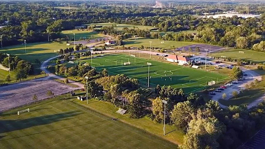 Aerial view of a sports complex with multiple soccer fields, a baseball field, parking lots, and surrounding trees in a suburban area with green rolling hills in the background.
