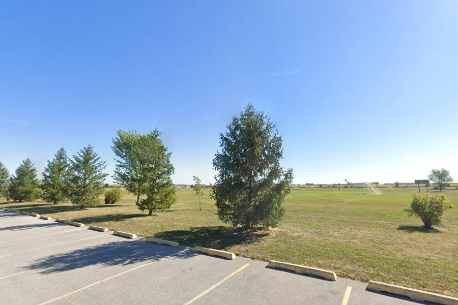 Empty parking lot adjacent to a grassy area with trees under a clear blue sky.