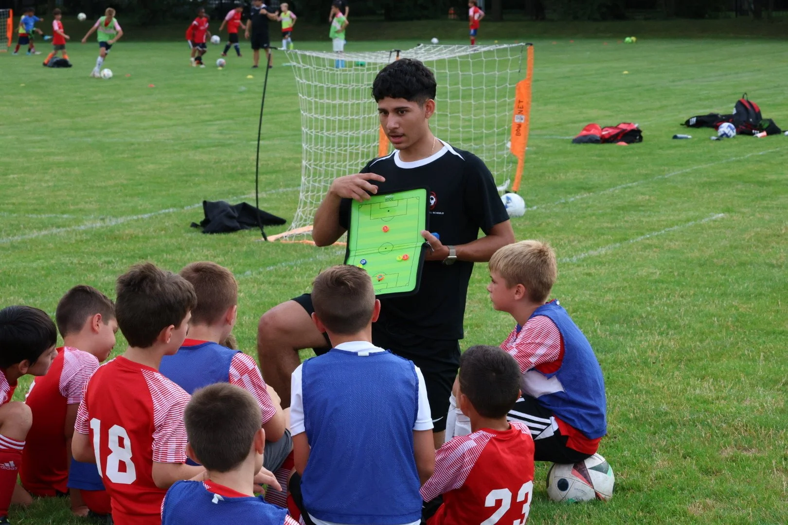 Soccer coach giving tactical instruction to young players during a practice session on a grassy field, with soccer goals and backpacks in the background.