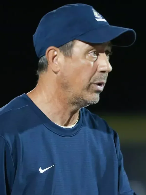 A middle-aged man wearing a navy blue baseball cap and matching Nike sports shirt with a white swoosh logo. He has short, salt-and-pepper hair and a beard. The background is dark and blurred.